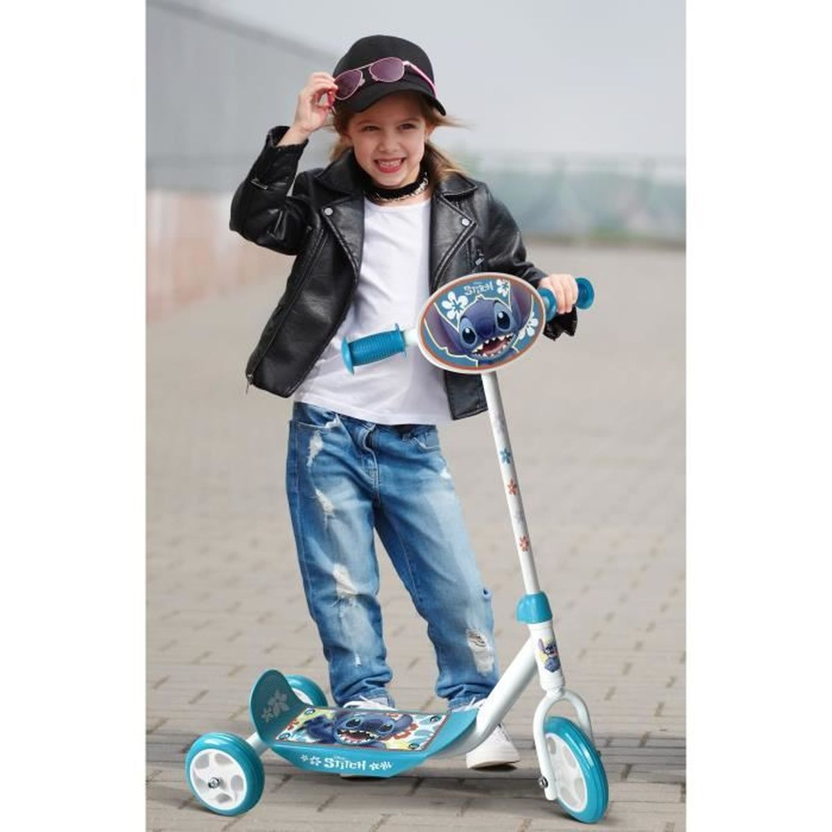 Una niña sonriente con chaqueta de cuero y jeans posa con un monopatín de Stitch azul y blanco.