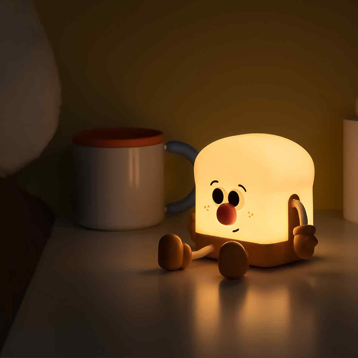 Eine süße, leuchtende Toasterlampe mit Gesicht sitzt auf einem weißen Tisch, im Hintergrund steht eine Tasse.