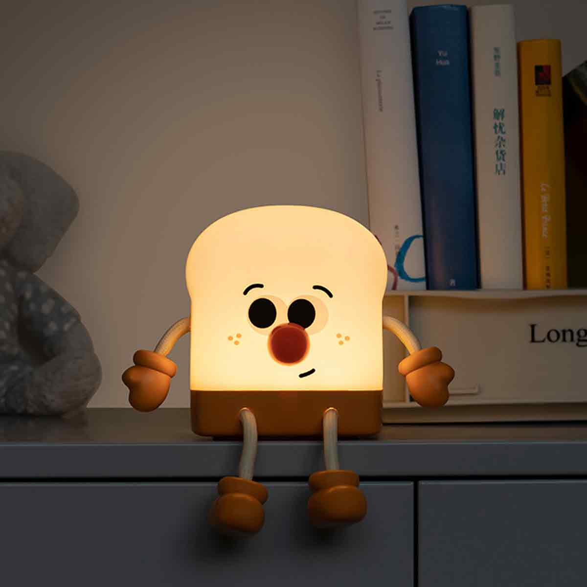 Eine niedliche, leuchtende Toast-Lampe mit Armen und Beinen sitzt auf einer grauen Oberfläche vor einem Bücherregal.