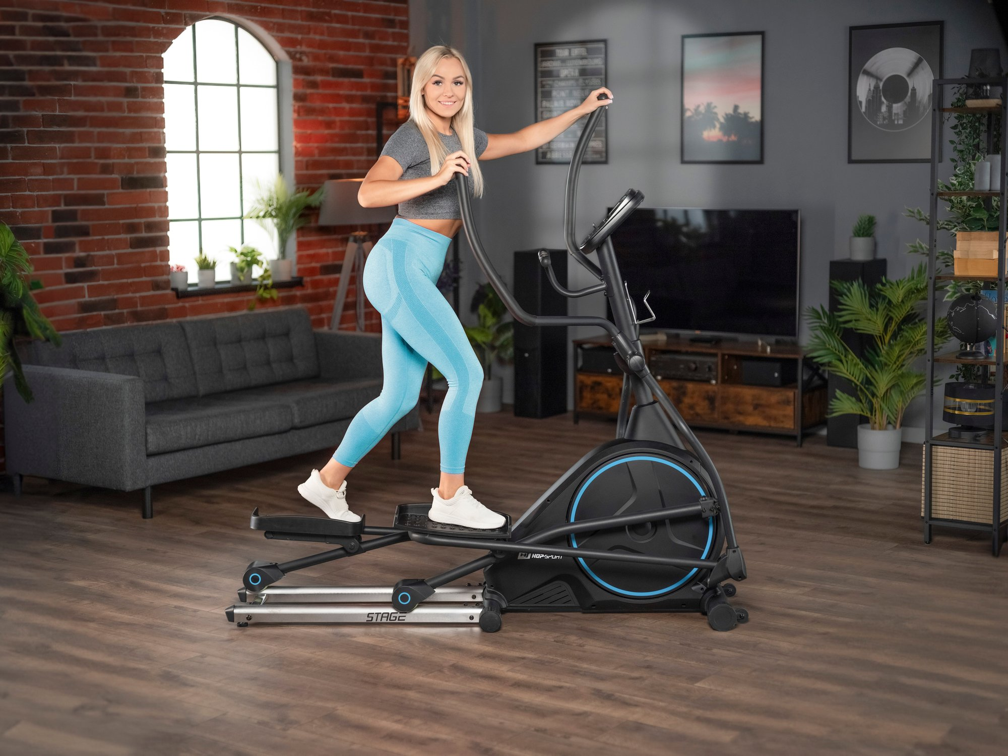 Eine blonde Frau in Sportkleidung trainiert zu Hause auf einem schwarzen Crosstrainer in einem modernen Wohnzimmer.