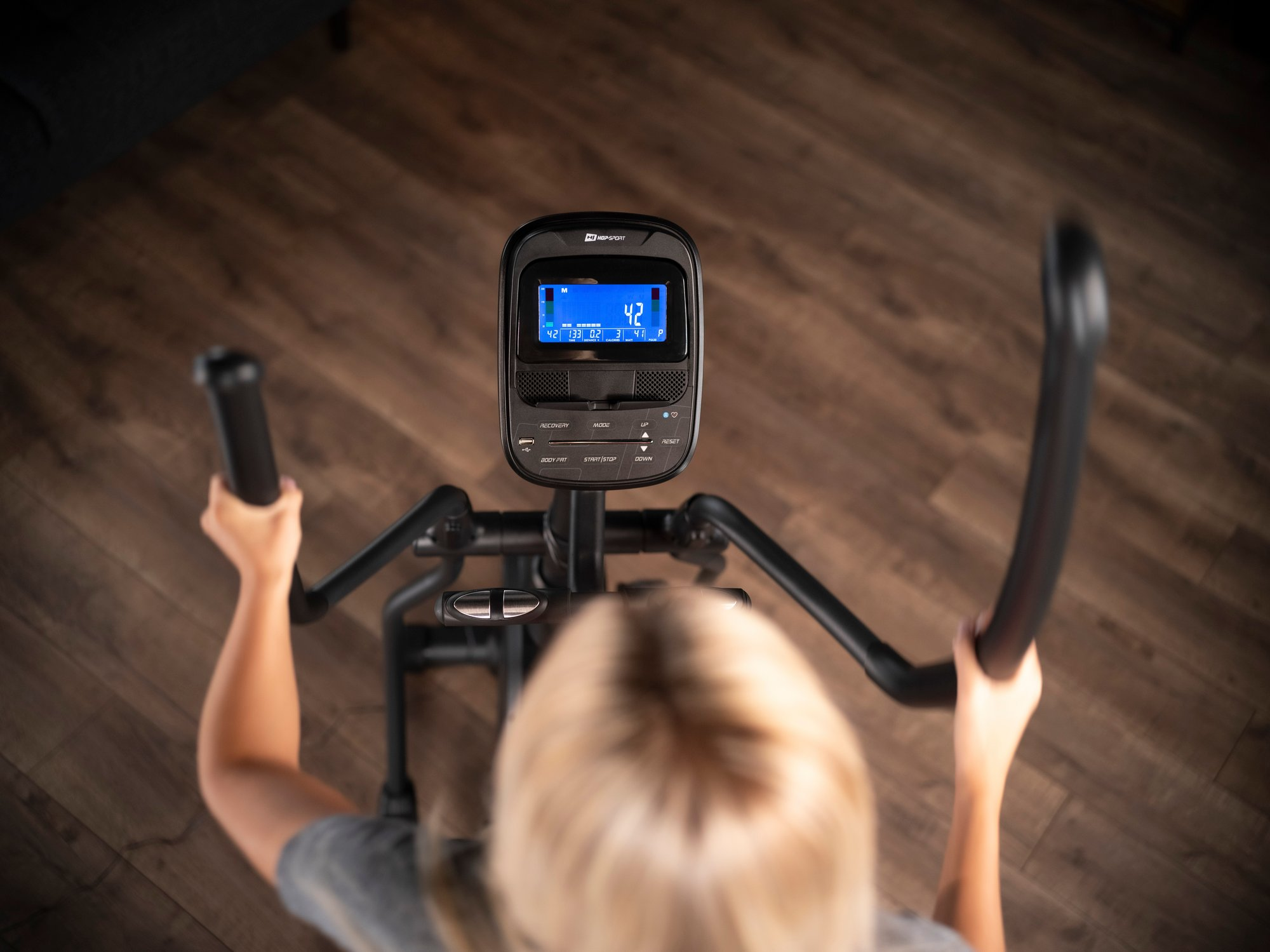 Top-down-Ansicht einer blonde Frau beim Training auf einem Crosstrainer auf Holzboeden.