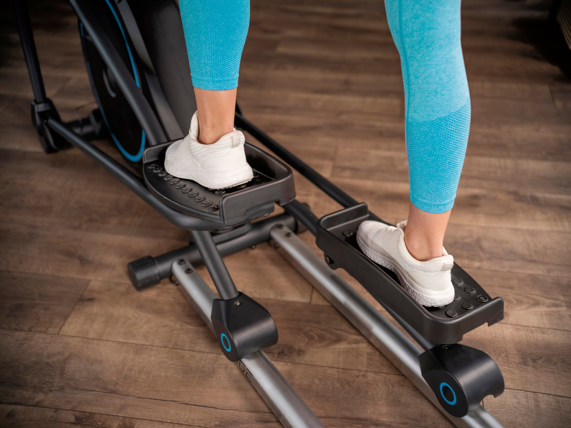 Person in blauen Leggings und weißen Sneakers trainiert auf einem schwarzen Heimtrainer.
