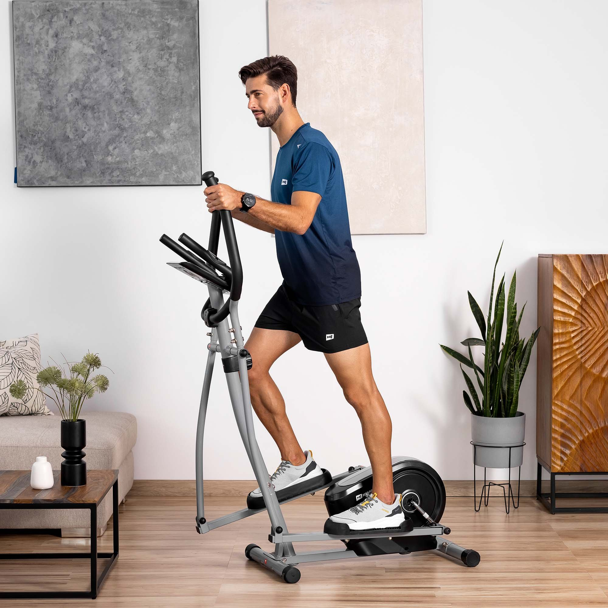 Ein Mann in Sportkleidung trainiert auf einem grauen Crosstrainer in einem hellen Wohnzimmer mit Wandkunst und Pflanzen.
