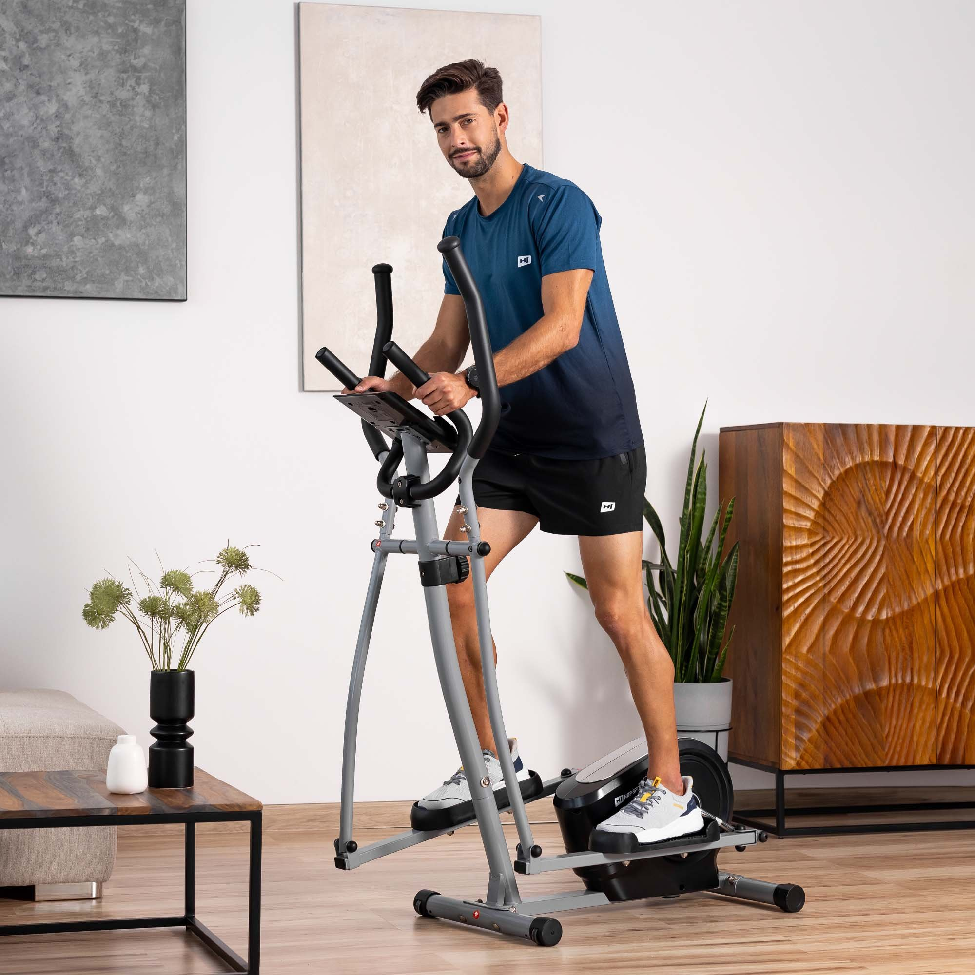 Ein Mann in blauem Shirt und schwarzen Shorts trainiert auf einem grau-schwarzen Crosstrainer in einem hellen Raum.