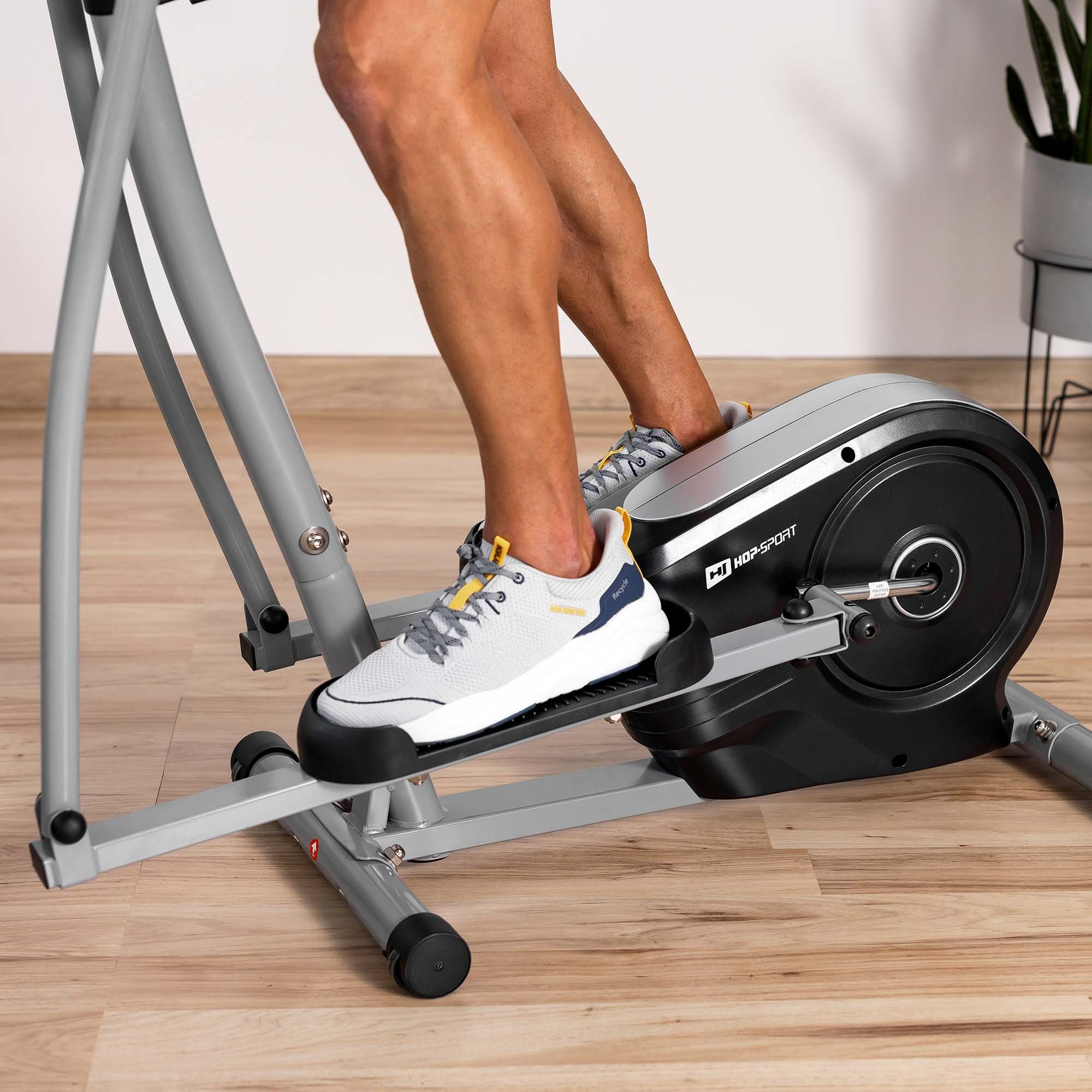 Person in weißem Sneaker trainiert auf einem grauen Crosstrainer in einem Raum mit Holzboden.