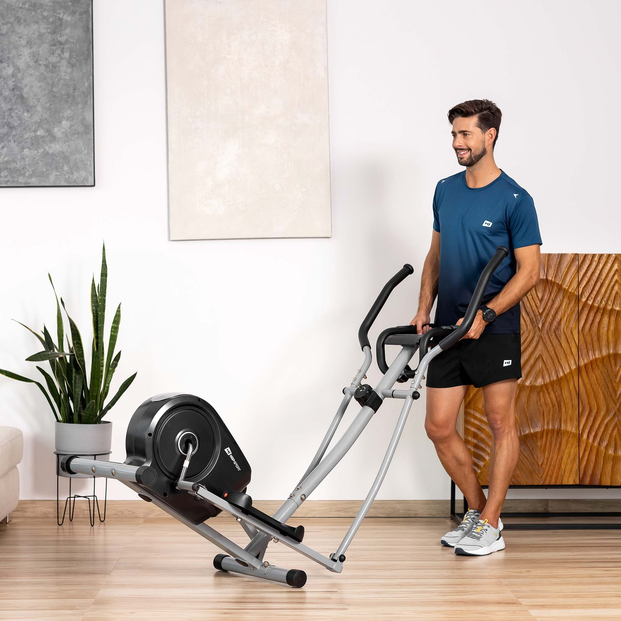 Ein Mann in Sportkleidung trainiert auf einem Crosstrainer in einem hellen Raum mit Pflanze und Kunstwerk.