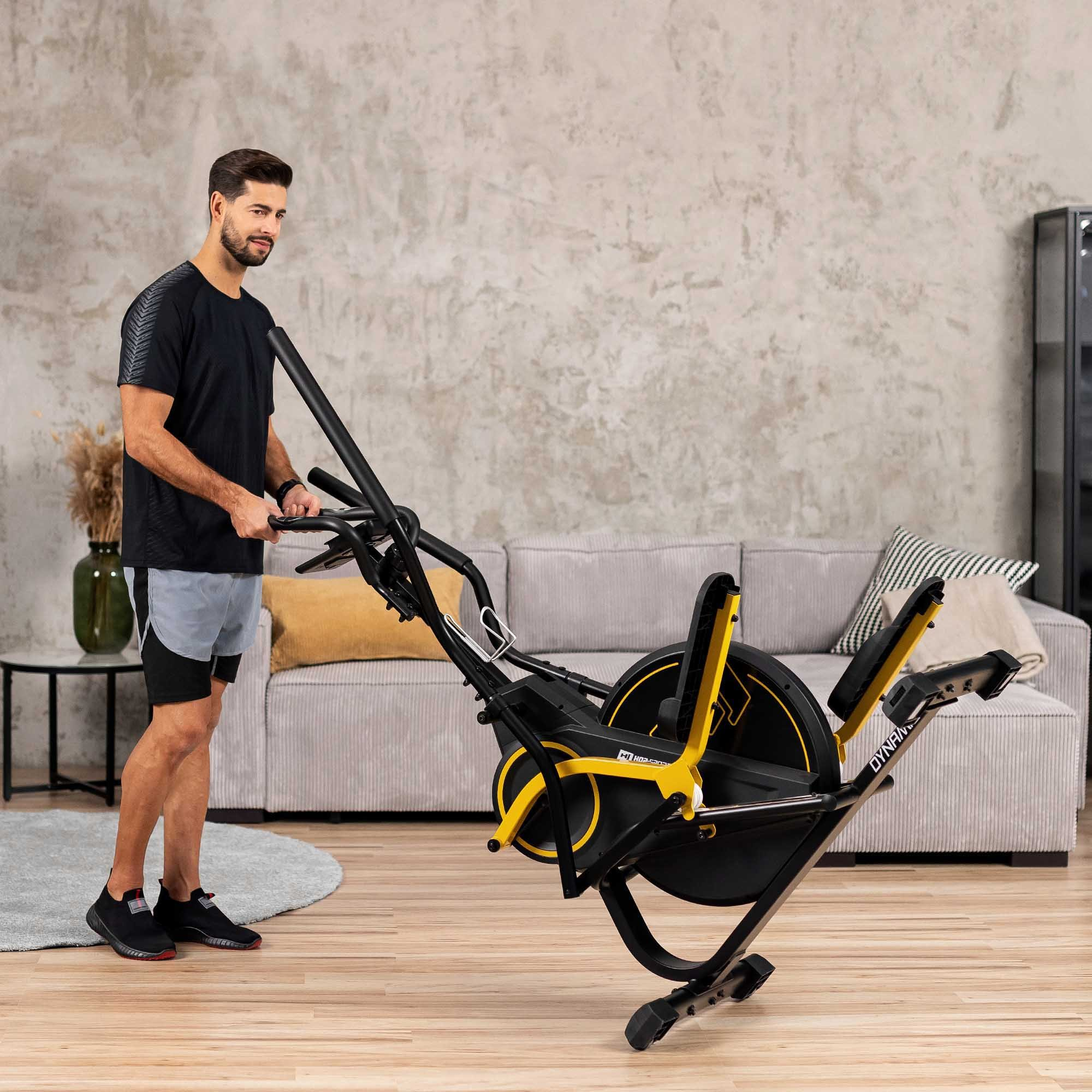 Ein Mann im schwarzen T-Shirt und grauen Shorts steht im Wohnzimmer und hält einen Crosstrainer.