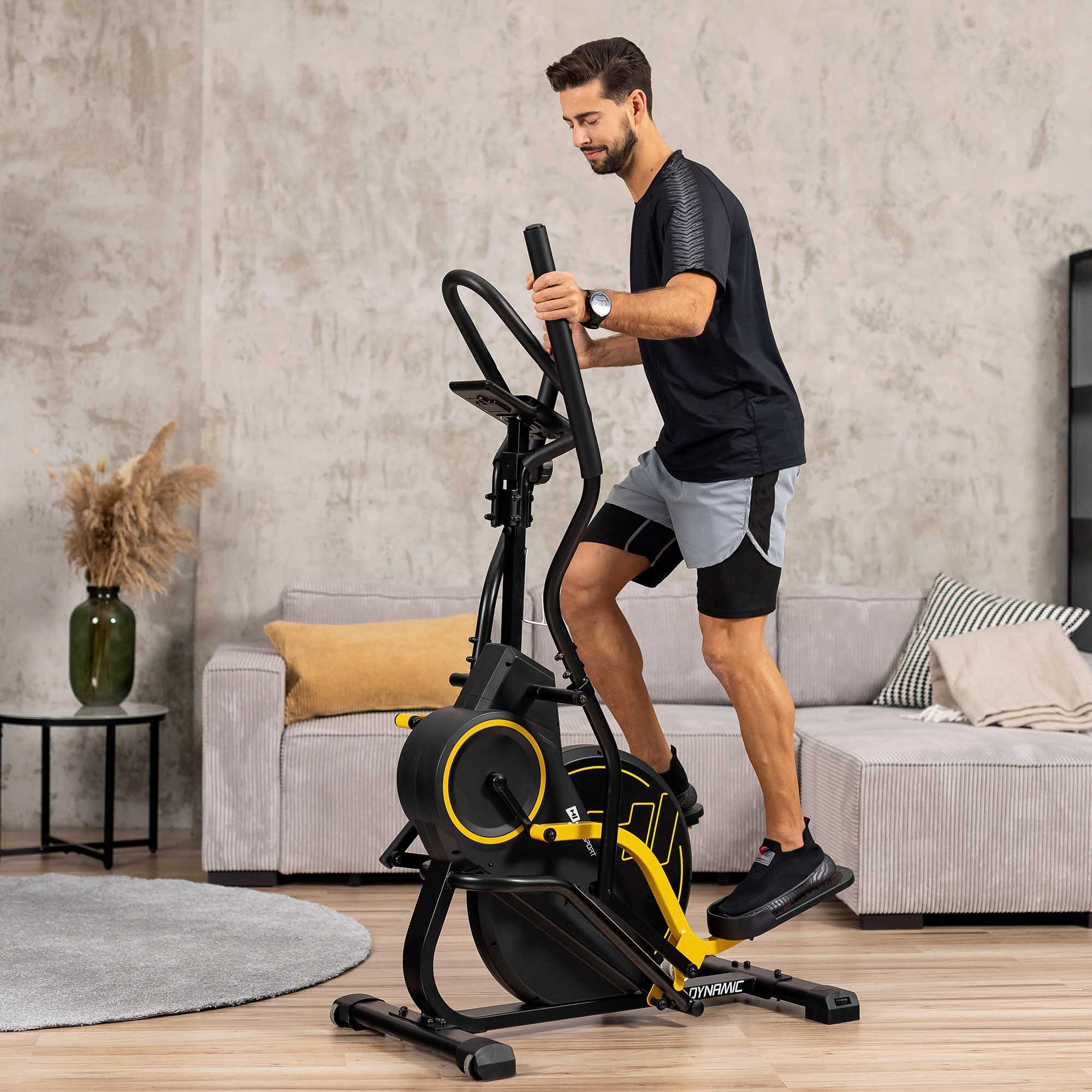 Ein Mann trainiert im Wohnzimmer auf einem schwarzen Crosstrainer mit gelben Akzenten.