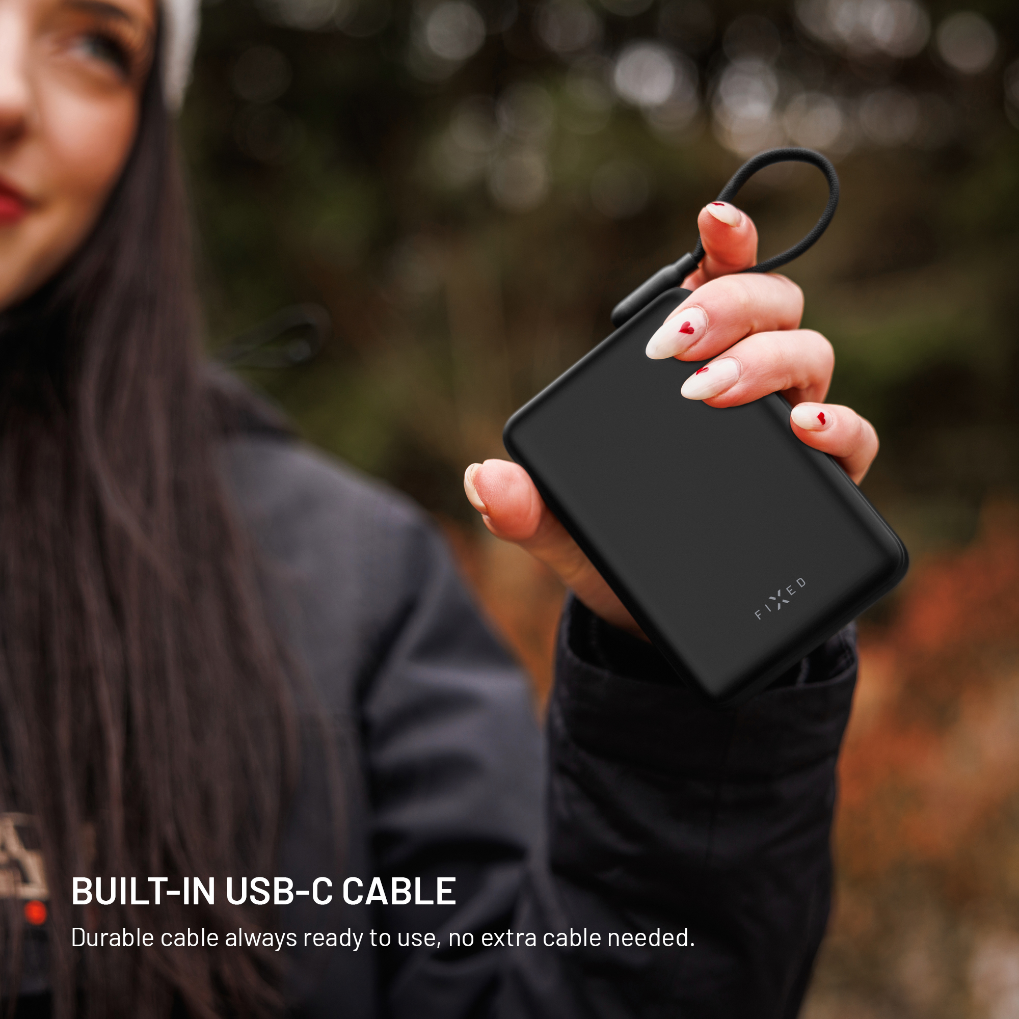 Eine Person hält eine schwarze Powerbank mit integriertem Kabel vor einen unscharfen Hintergrund.