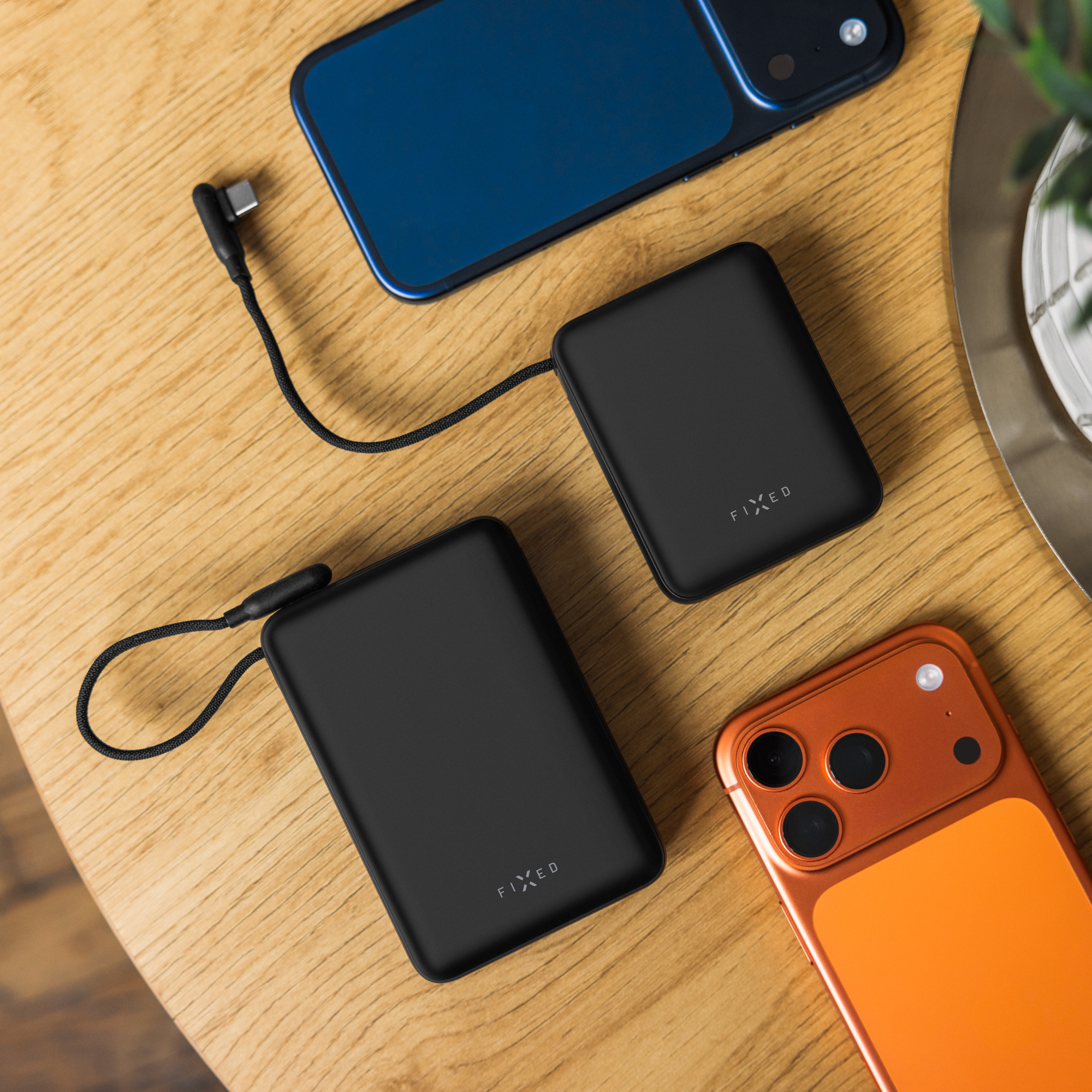 Zwei schwarze Fixed Powerbanks mit Kabeln auf Holzoberfläche neben blauen und orangen Smartphones.