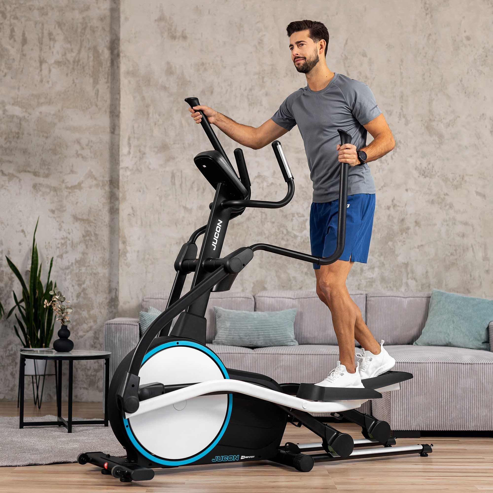 Ein Mann trainiert im Wohnzimmer auf einem schwarzen Crosstrainer vor einem Sofa und einer gemauerten Wand.