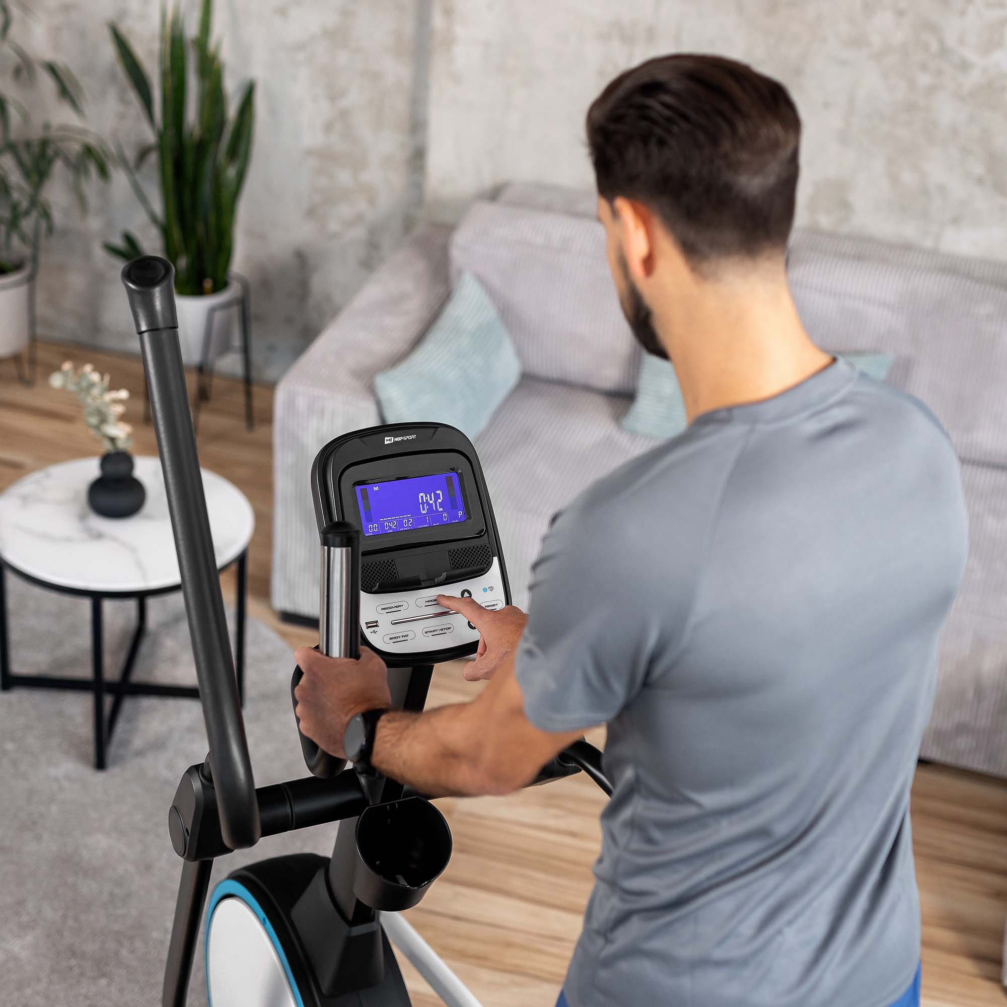 Ein Mann in grauem Shirt trainiert zu Hause auf einem Crosstrainer in einem modernen Wohnzimmer.