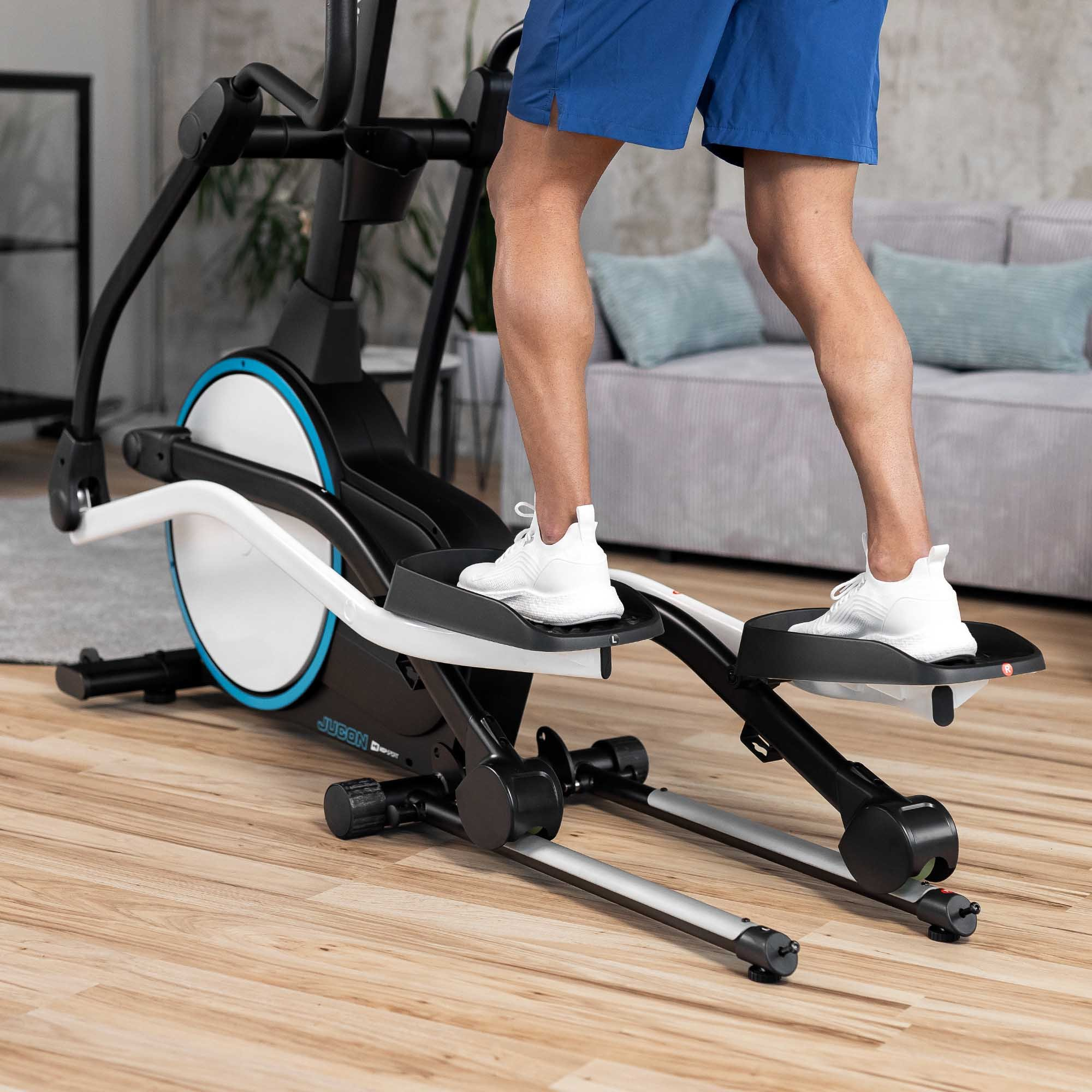 Person in blauen Shorts und weißen Sneakers trainiert auf einem Crosstrainer auf Holzboden vor einem Sofa.