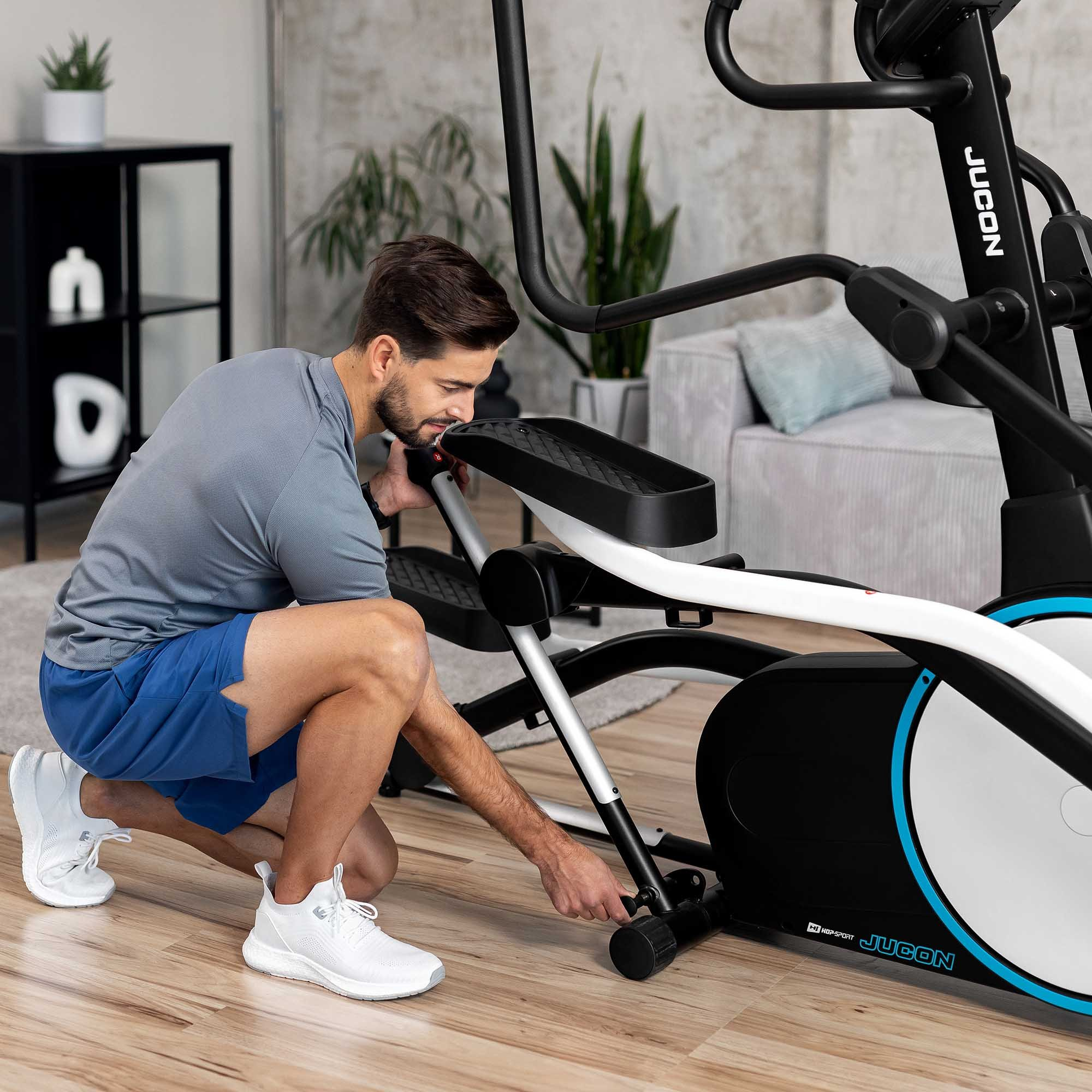 Ein Mann im grauen Hemd und blauen Shorts kniet und stellt einen schwarz-weißen Crosstrainer ein.