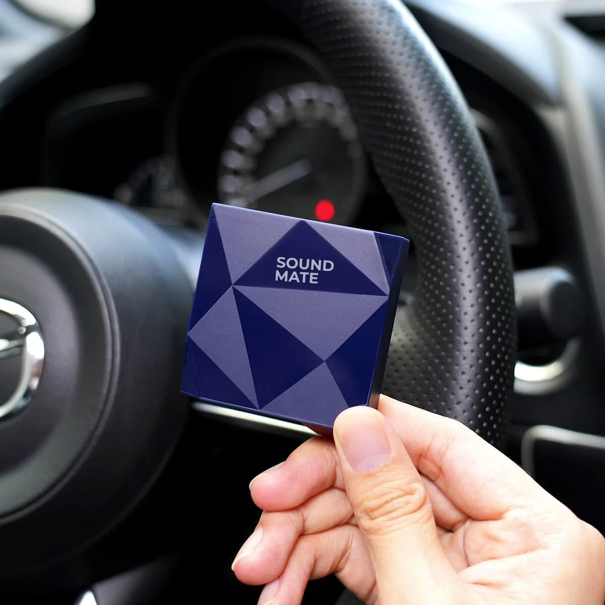 Eine Hand hält eine blaue Sound Mate Box vor einem Auto-Lenkrad.