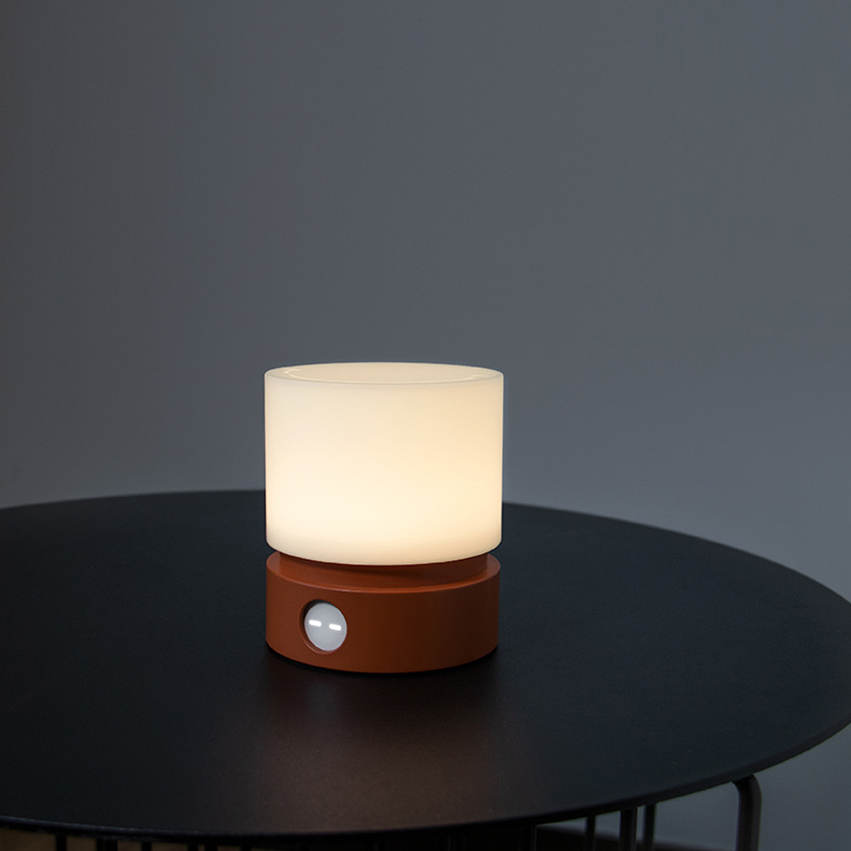 Eine kleine beleuchtete Lampe mit einem orangen Sockel steht auf einem schwarzen Tisch vor einer grauen Wand.