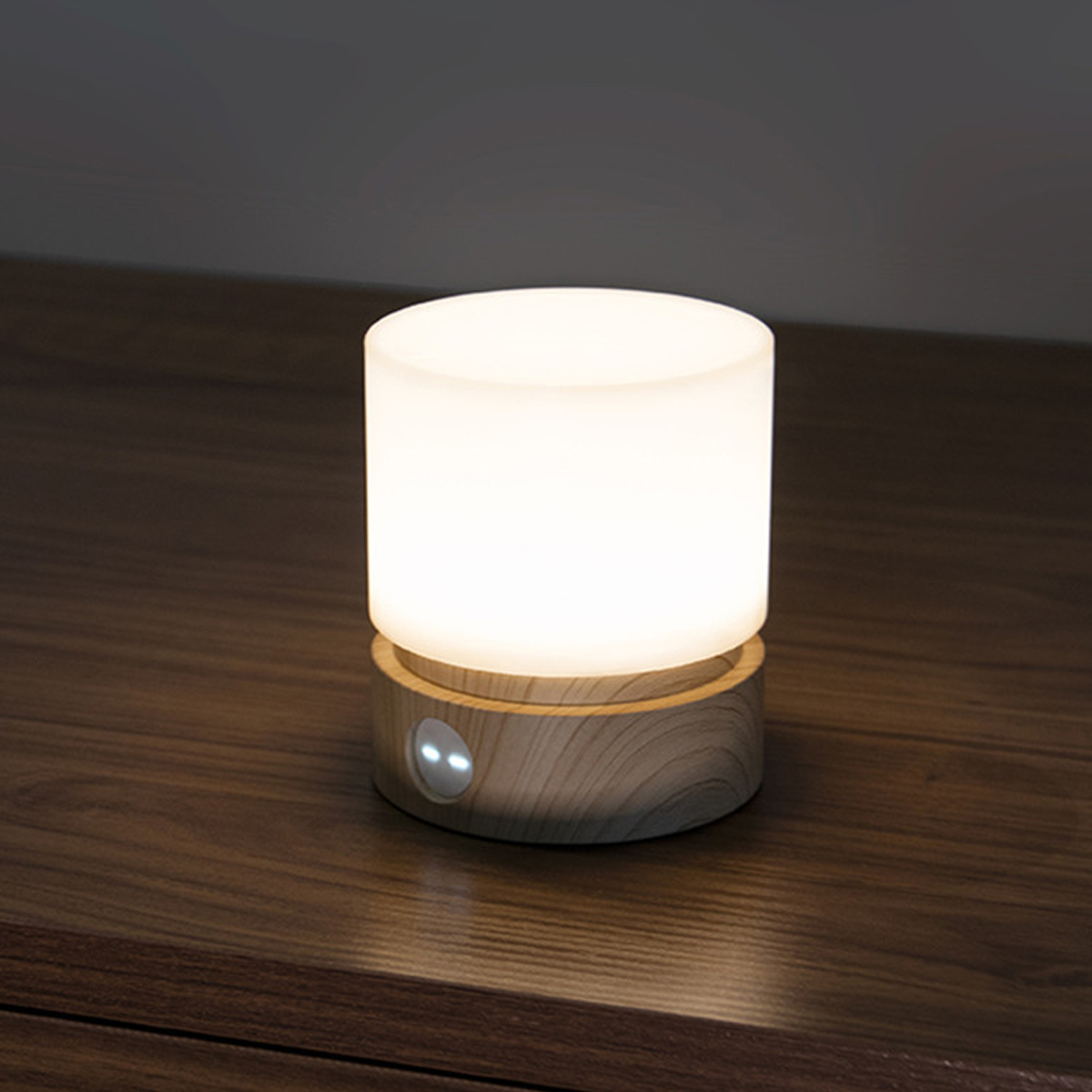 Eine leuchtende zylindrische Nachttischlampe mit Holzsockel auf einem dunklen Holztisch vor einer dunklen Wand.