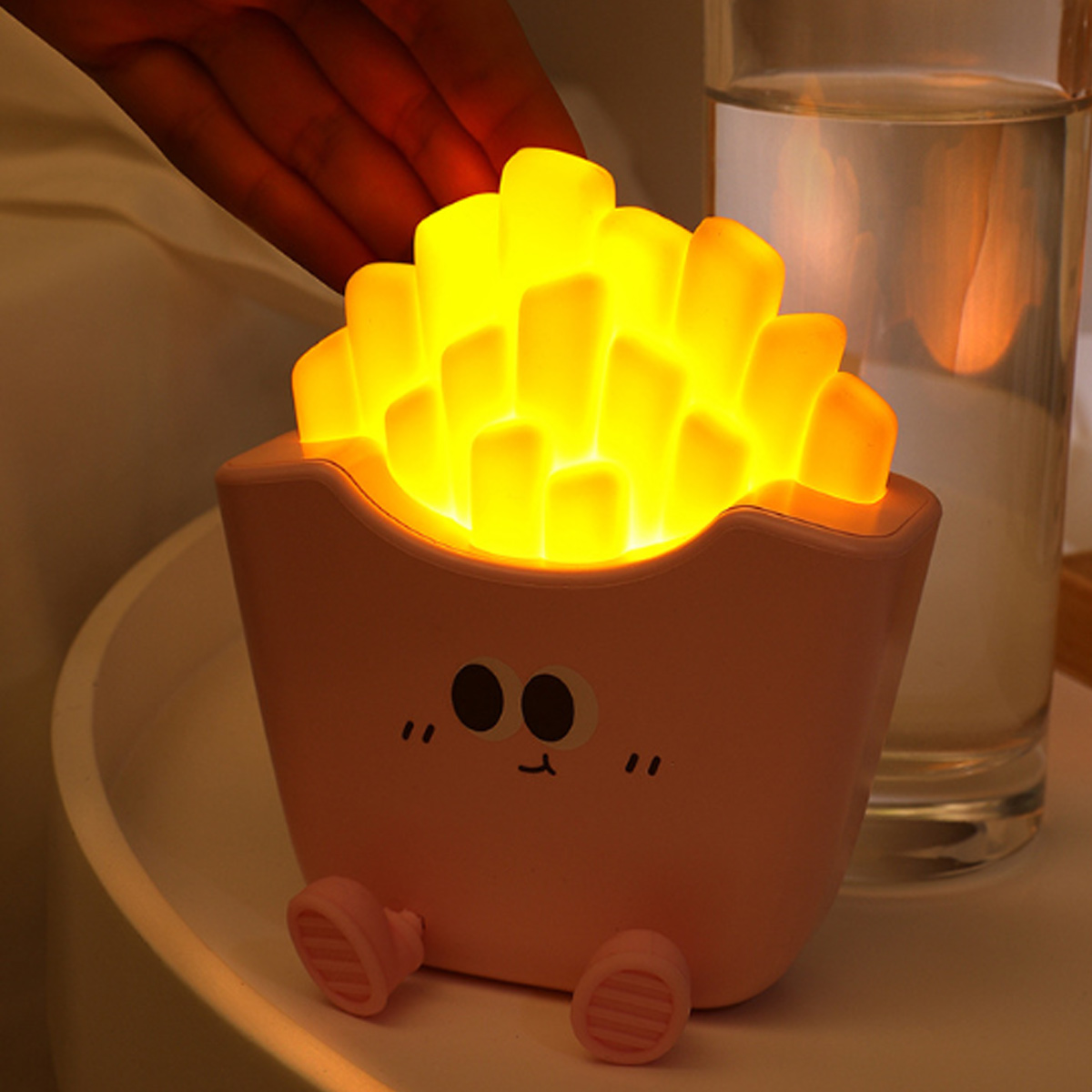 Eine rosafarbene Nachtlampe im Pommes-Design mit niedlichem Gesicht leuchtet gelb auf einem weißen Tisch.