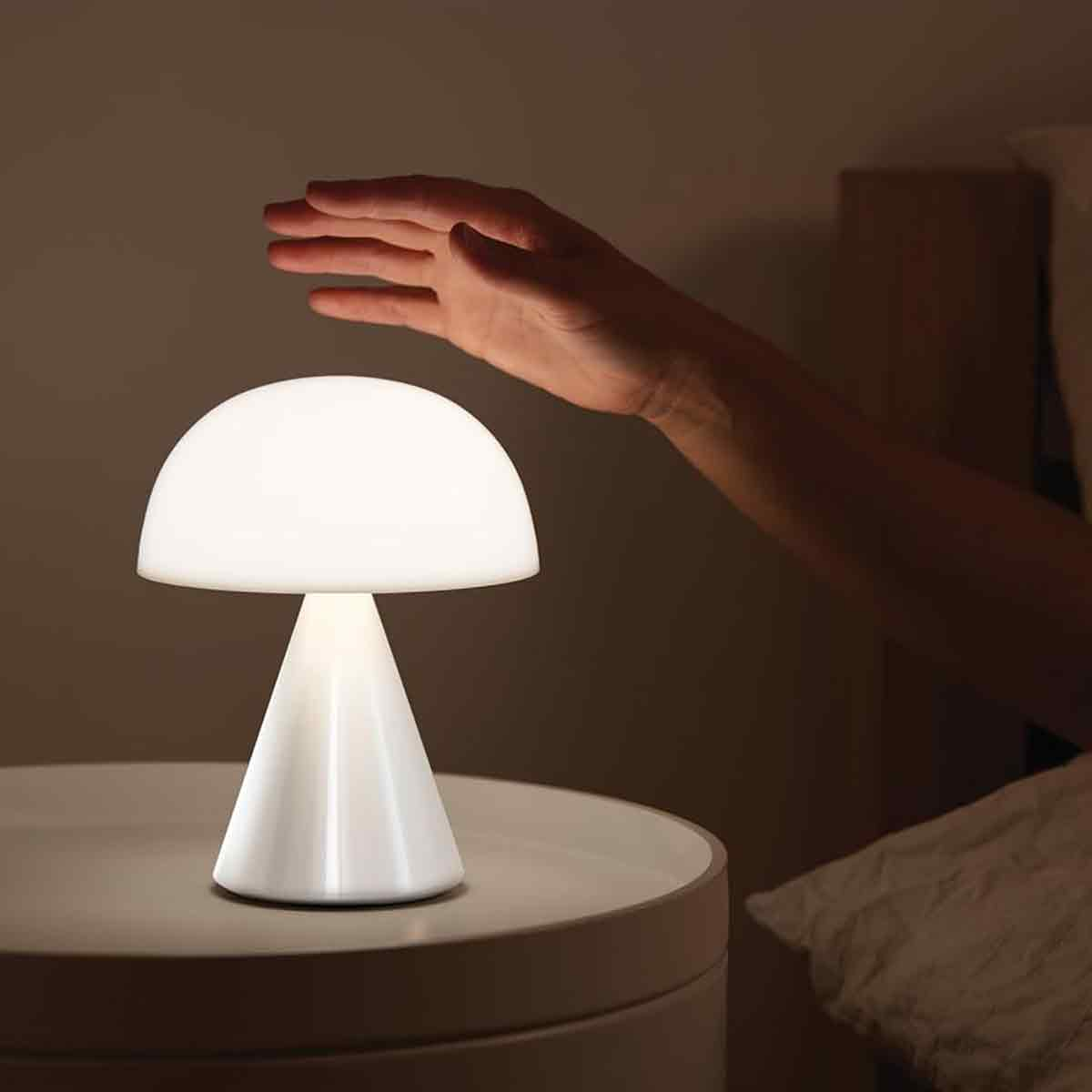 Eine Hand schwebt über einer kleinen, pilzförmigen weißen Lampe auf einem Nachttisch.