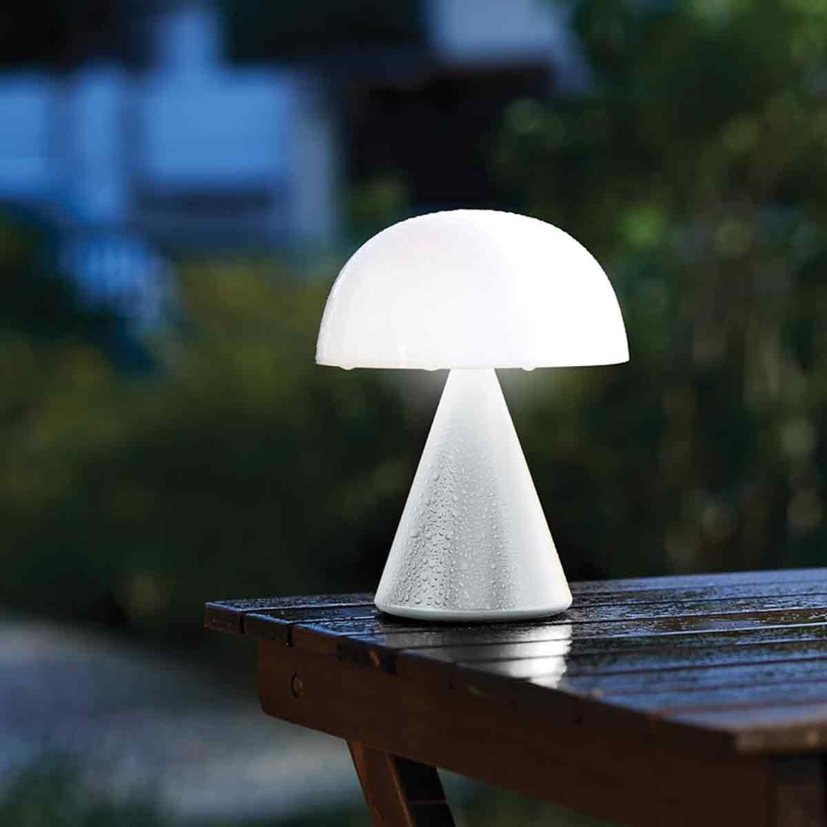 Eine weiße Pilzlampe steht auf einem nassen Holzschreibtisch im Freien bei Dämmerung.