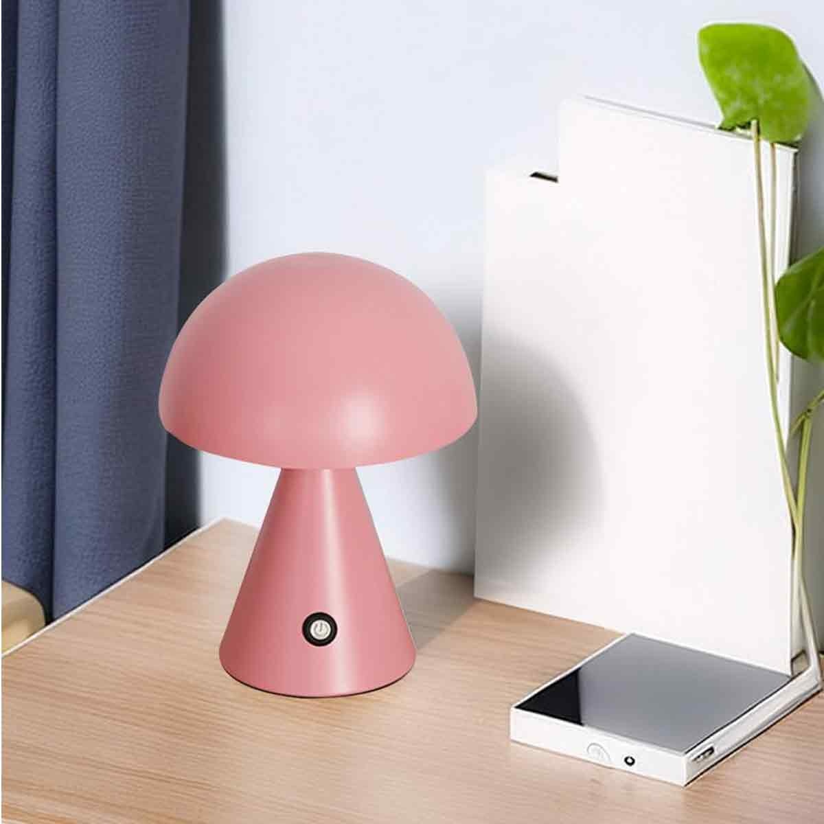 Eine rosa pilzförmige Schreibtischlampe mit Ein/Aus-Schalter steht auf einem Holztisch neben einem weißen Buch und einer Pflanze.