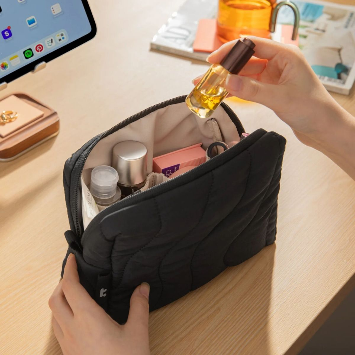 Eine Person legt eine kleine bernsteinfarbene Glasflasche in eine offene schwarze Kosmetiktasche auf einem Holztisch.