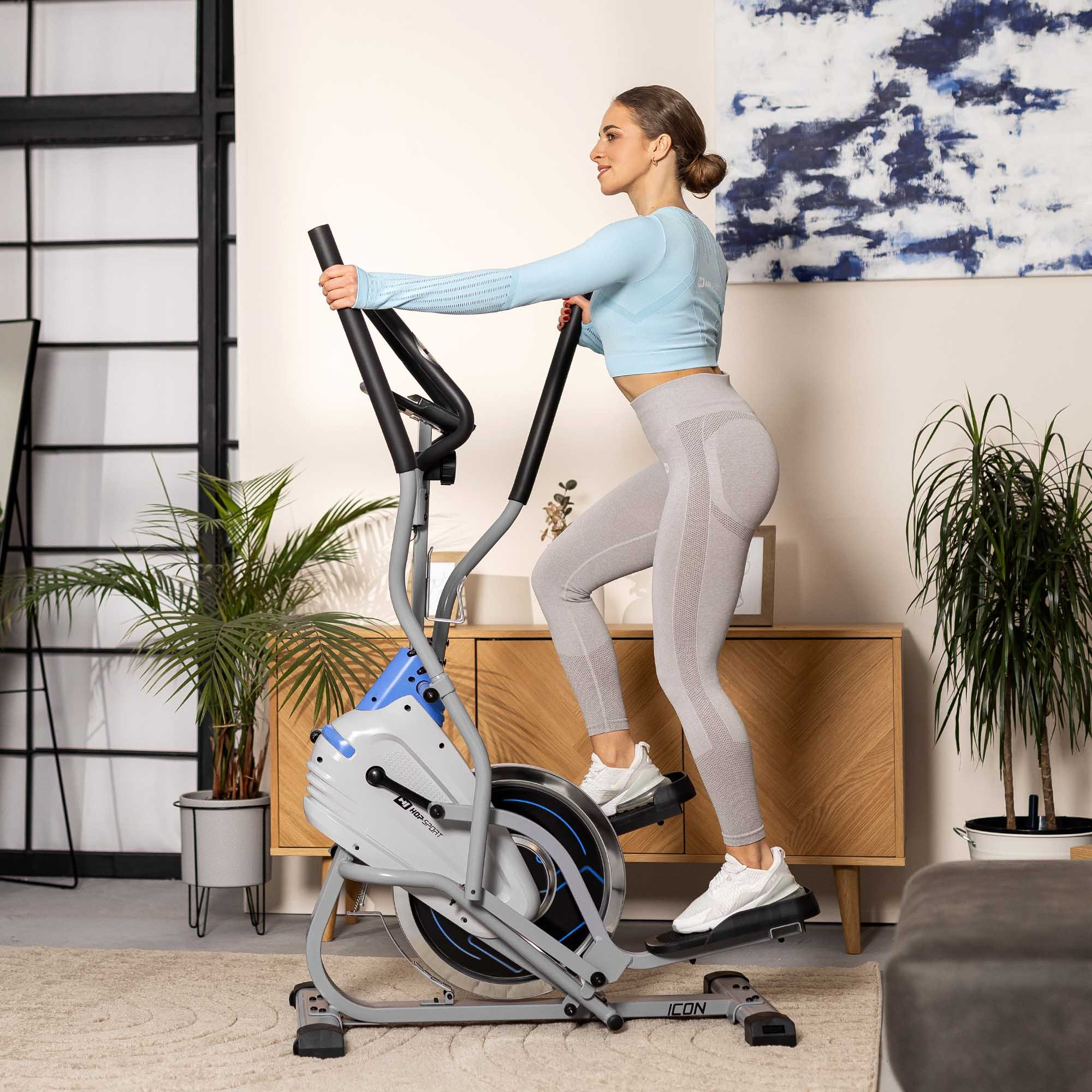 Eine Frau in Sportkleidung trainiert drinnen auf einem grauen Crosstrainer.