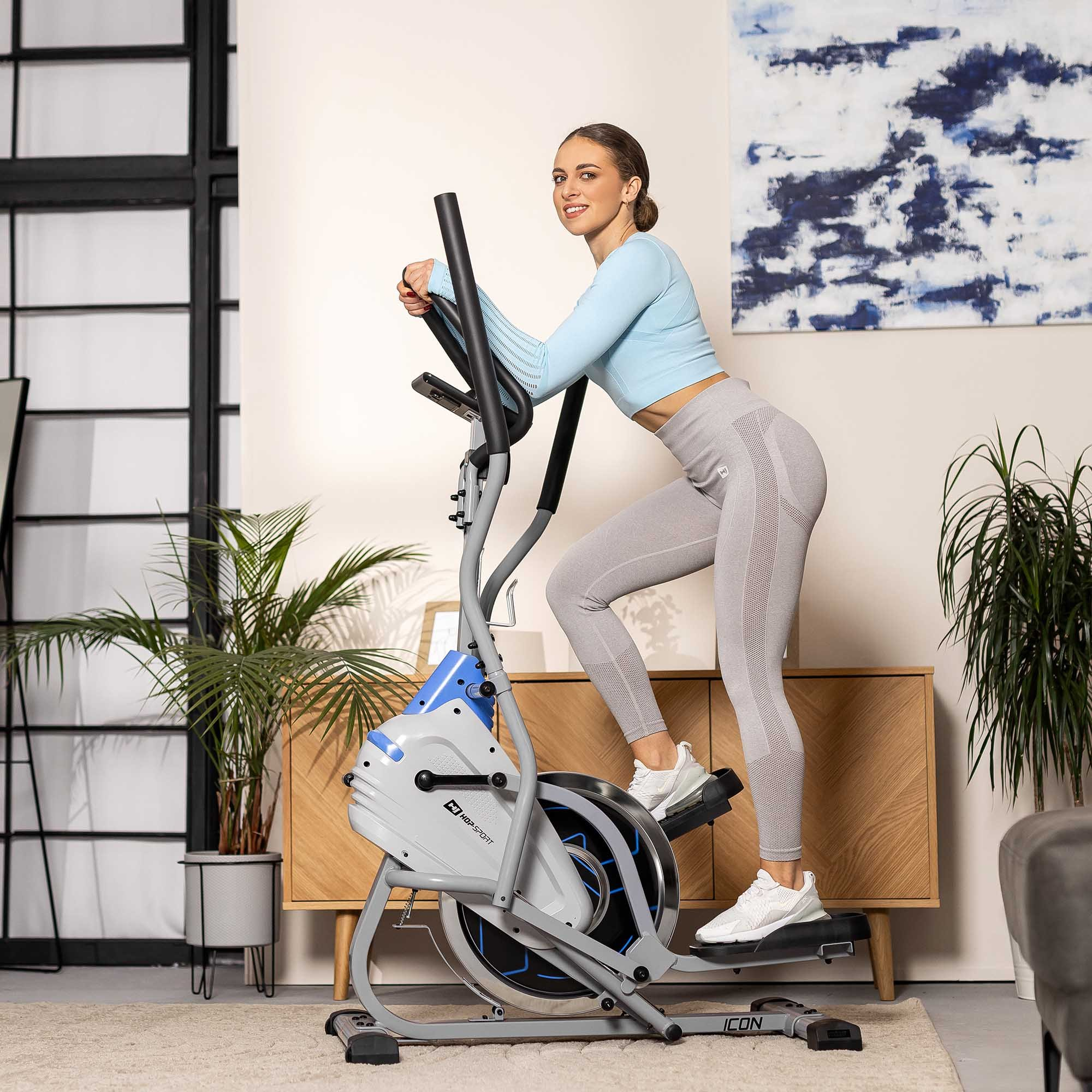 Eine Frau in Sportkleidung trainiert zu Hause auf einem grauen Crosstrainer.