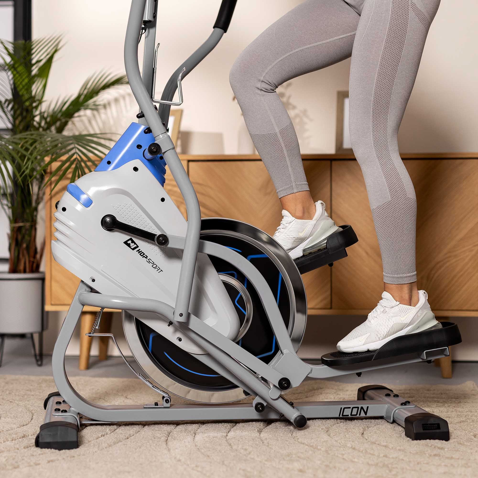 Eine Person in grauen Leggings nutzt einen graublauen Crosstrainer auf einem Teppich, mit Holzschrank im Hintergrund.
