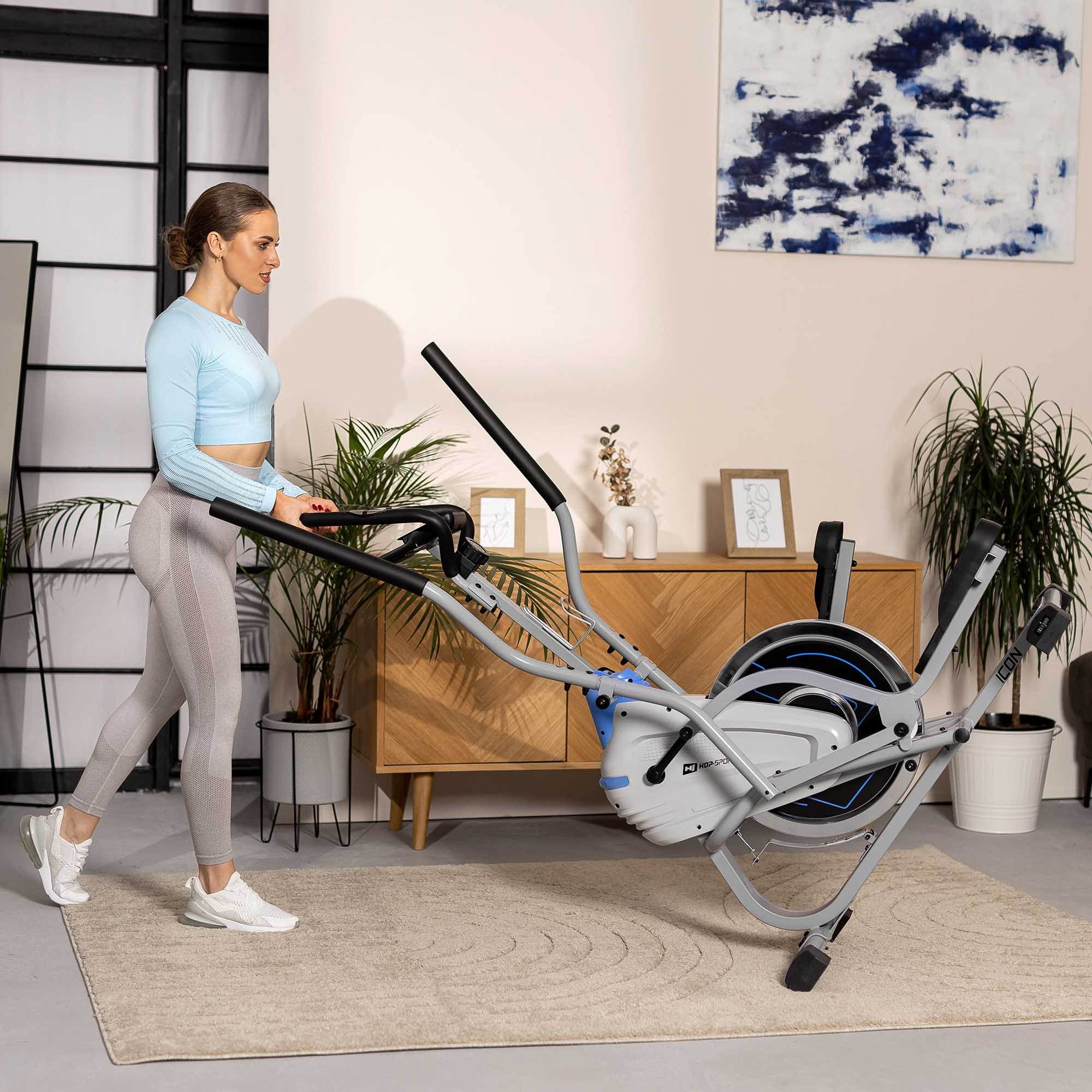 Eine Frau in Sportkleidung nutzt ein grau-blaues Crosstrainer-Gerät in einem hellen, modernen Raum.