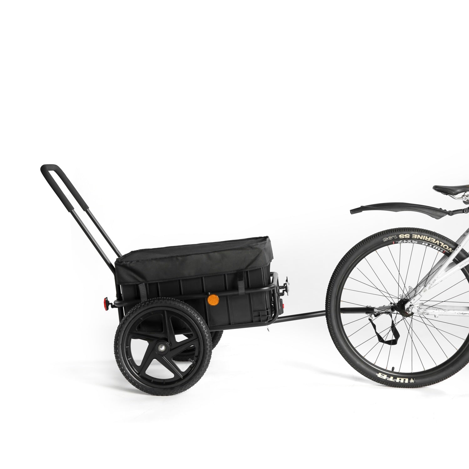 Un remolque de carga para bicicleta negro con ruedas y mango, acoplado a la rueda trasera de una bicicleta sobre fondo blanco.