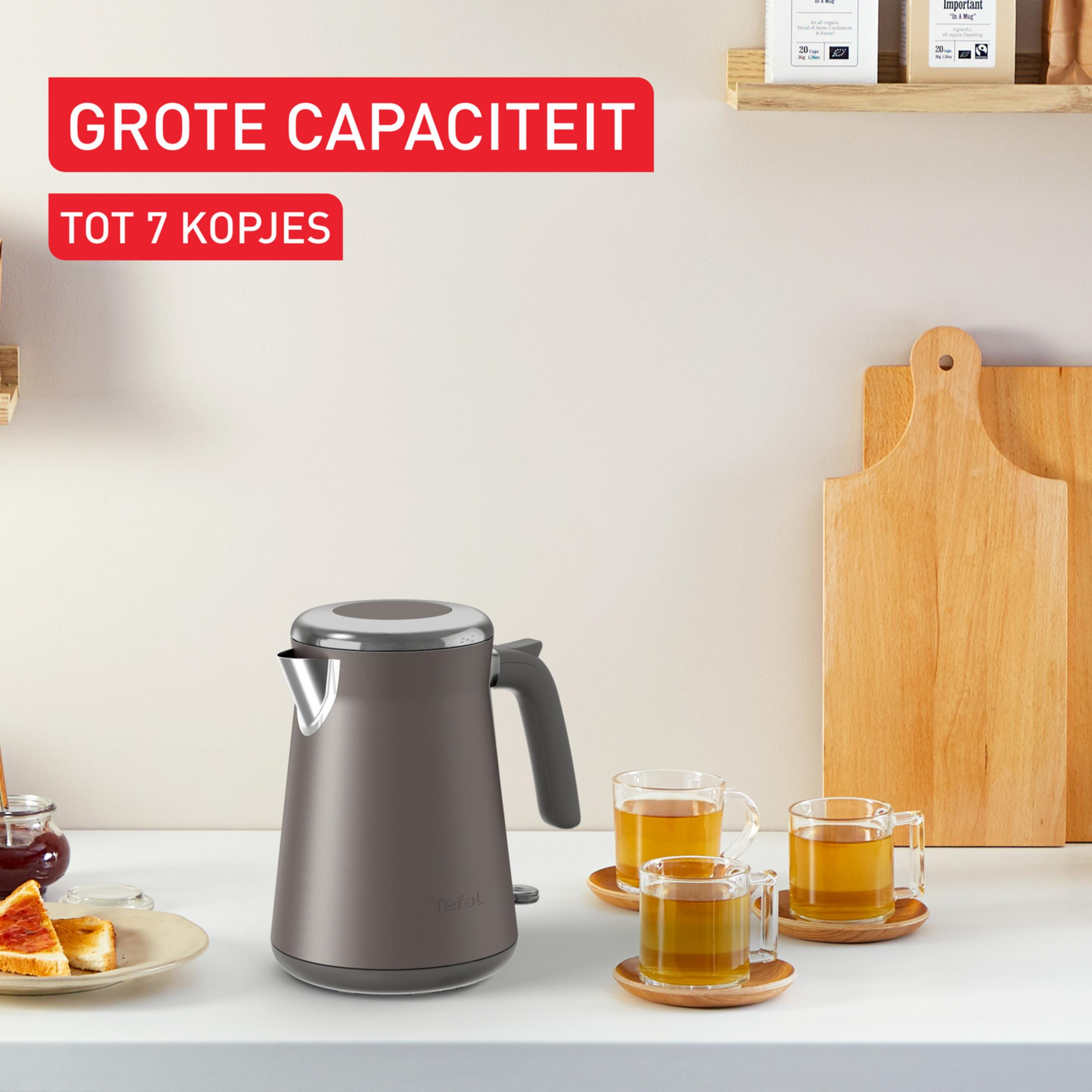 Bollitore elettrico grigio su un bancone con tre tazze di tè e un tagliere di legno sullo sfondo.