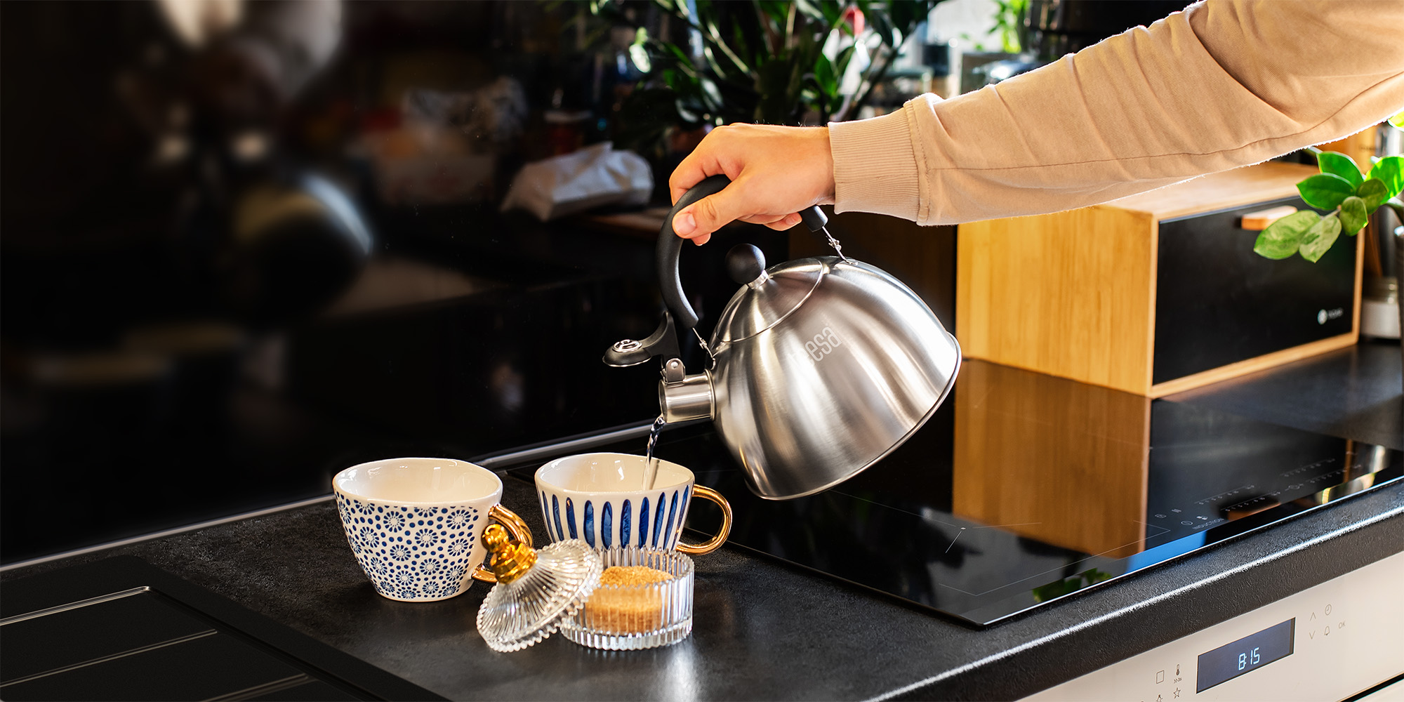 Una mano versa acqua da un bollitore argentato in una tazza su un piano cucina nero.
