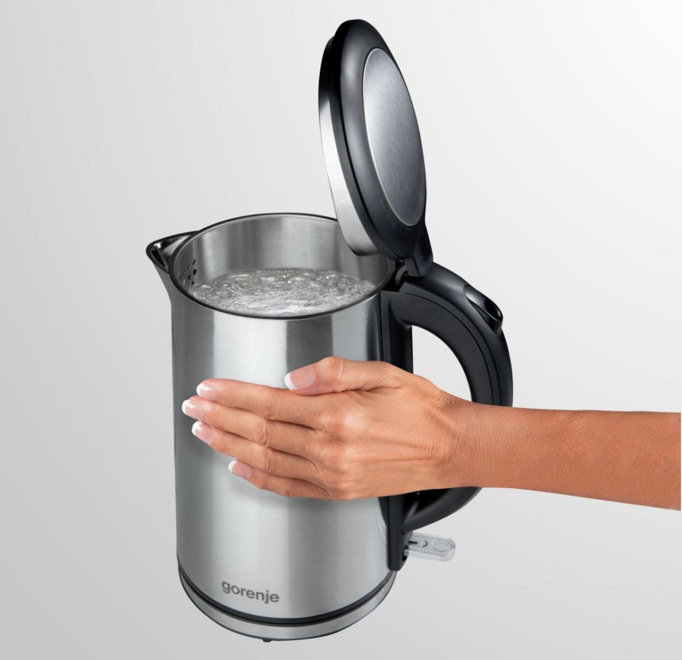 Mano che tocca un bollitore elettrico aperto con acqua bollente su sfondo grigio.