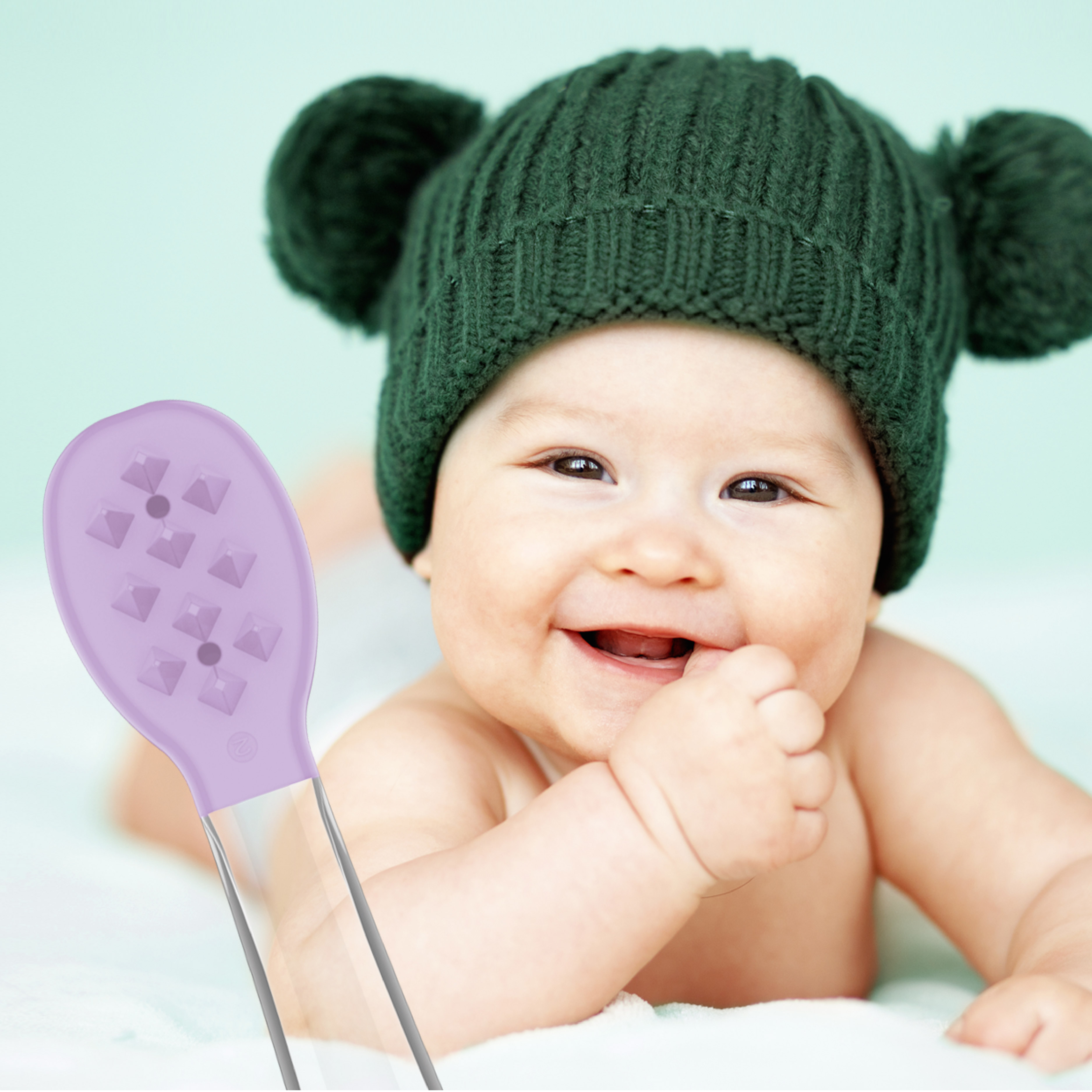 Ein lächelndes Baby mit grüner Mütze schaut auf eine lila Silikonbürste im Vordergrund.