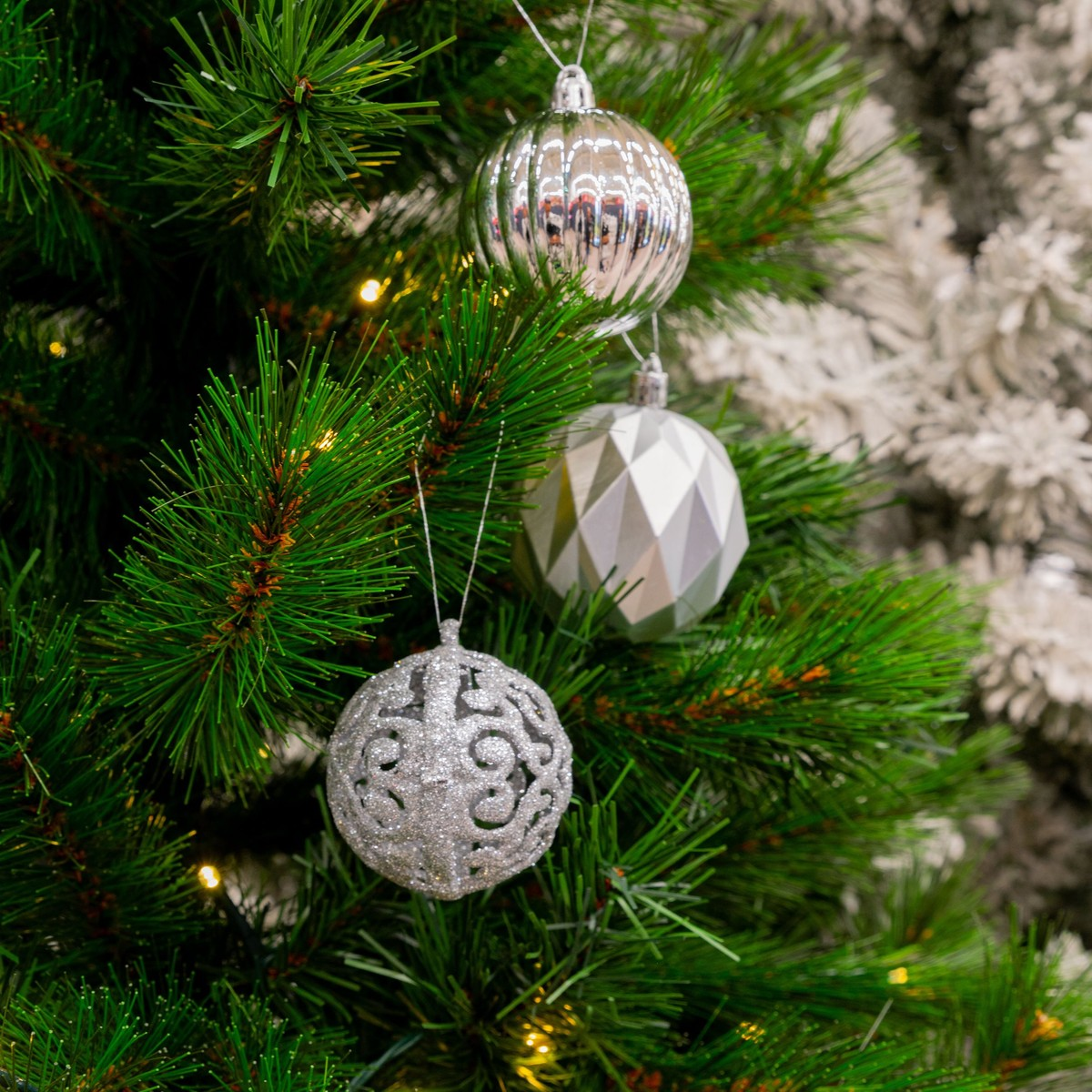 Three silver Christmas ornaments hang on a green artificial pine tree with soft lights.