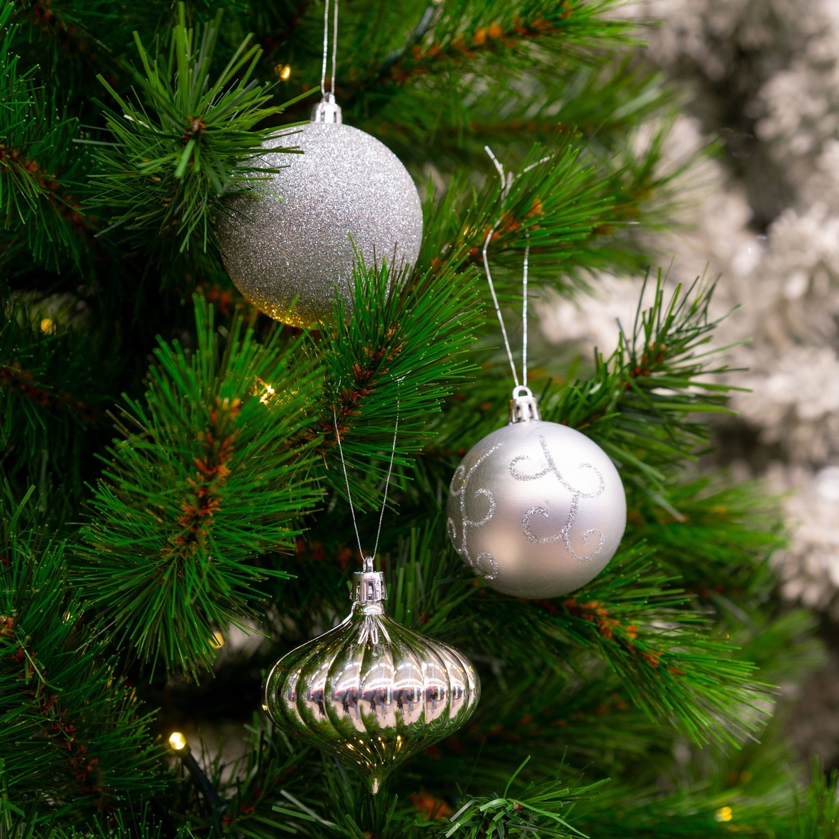 Three silver Christmas ornaments hang on a green pine tree with lights.