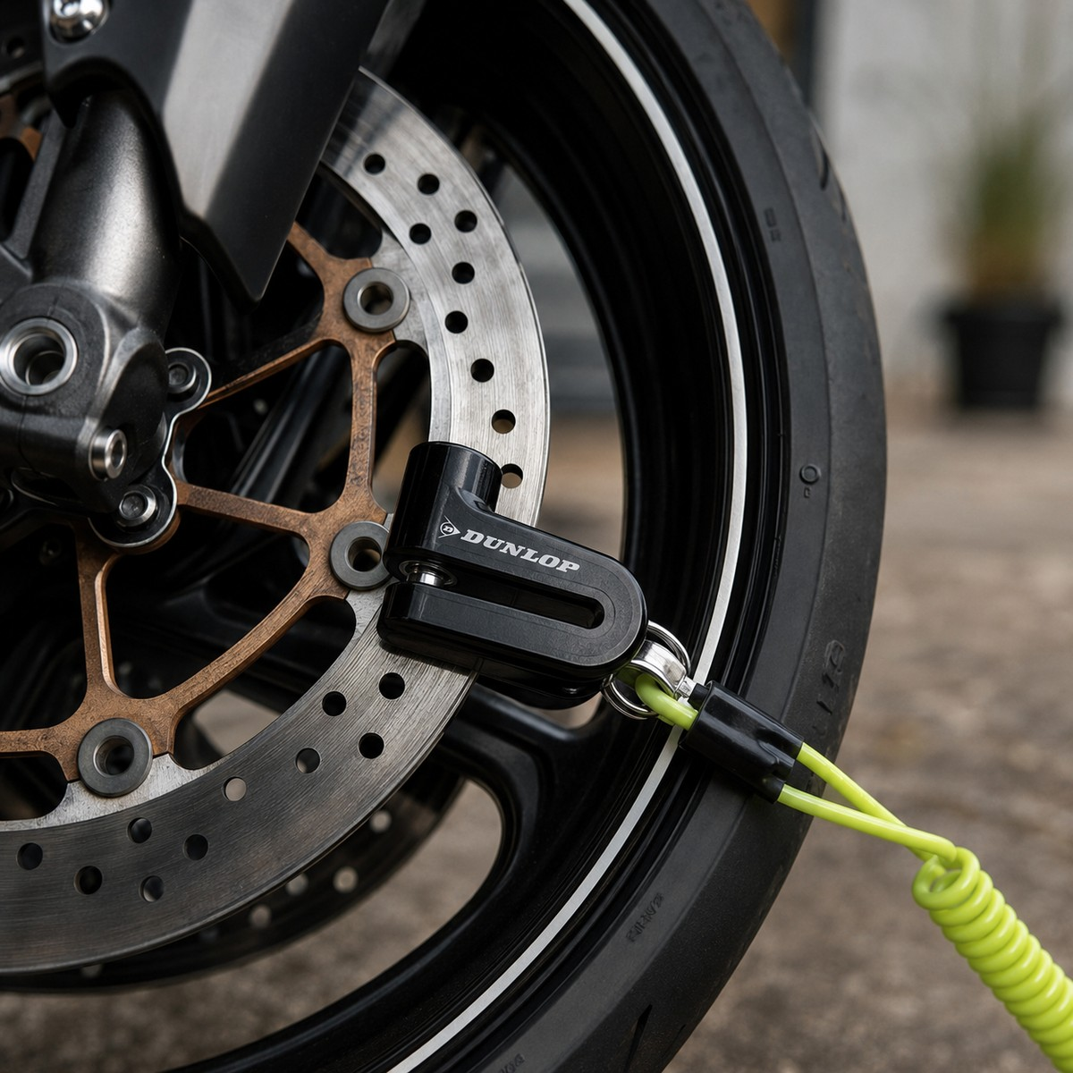 A black Dunlop disc lock is attached to a motorcycle's front brake rotor, connected to a neon green reminder cable.
