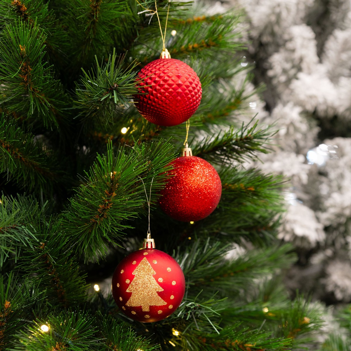 Three red Christmas baubles hang vertically on a green pine tree, with a snowy tree in the background.