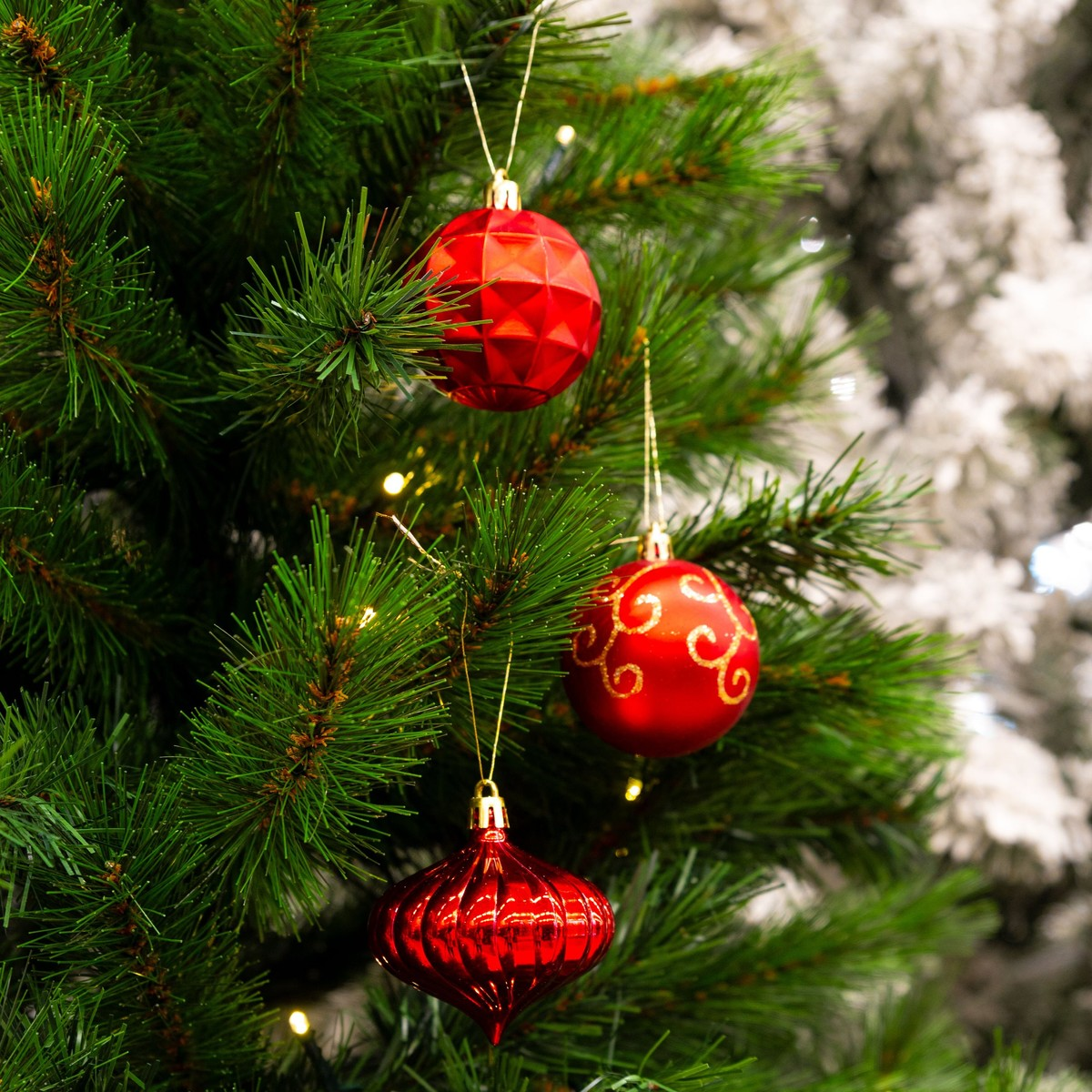 Three shiny red Christmas ornaments hang on a green artificial pine tree with soft white lights.