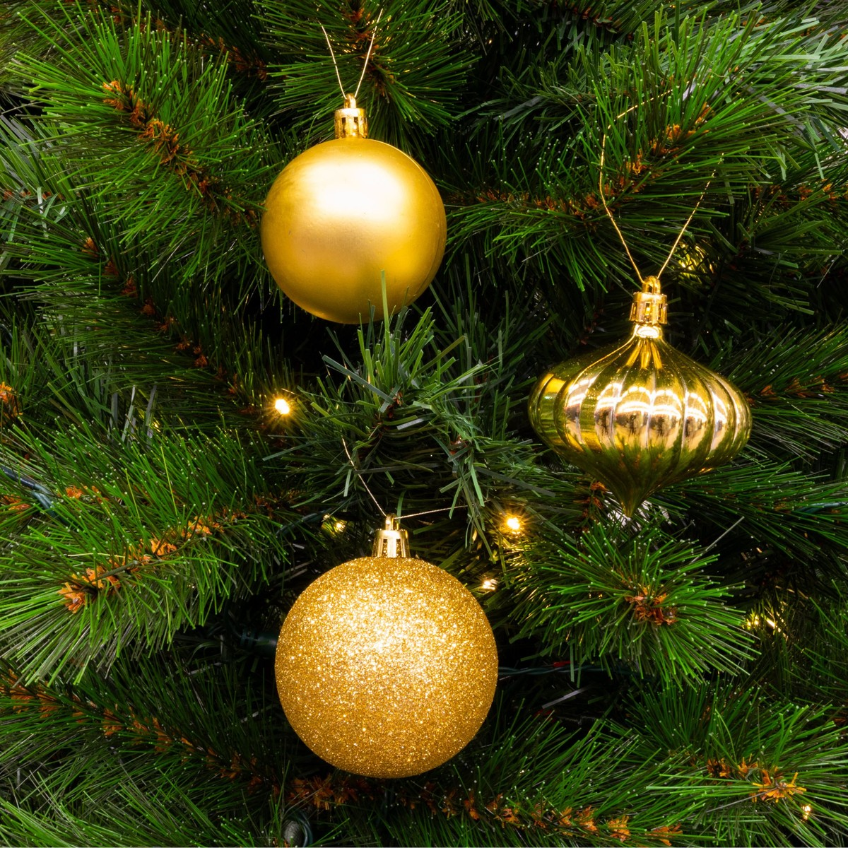 Three gold Christmas ornaments hang on a green Christmas tree with small lights.