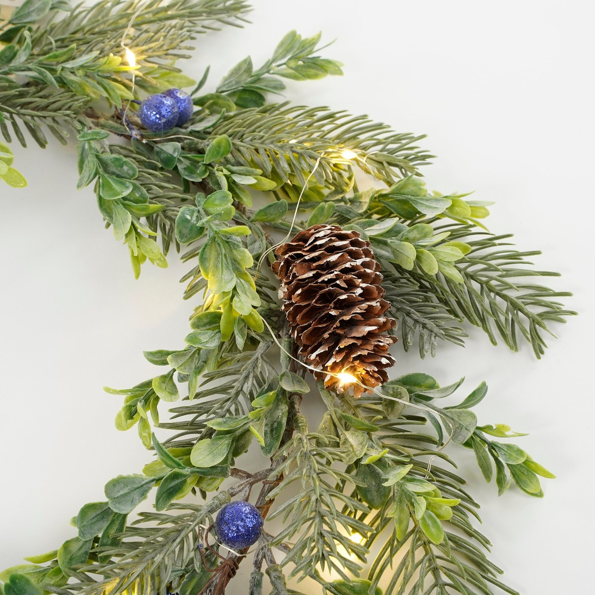 A festive wreath with green pine branches, a brown pinecone, blue berries, and glowing fairy lights on white.