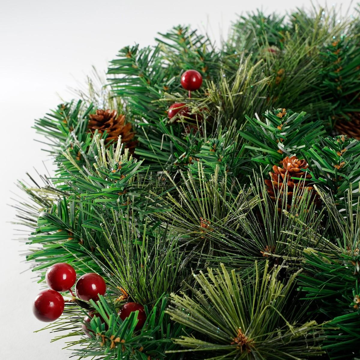 Artificial Christmas wreath with green branches, red berries, and pinecones on a light background.