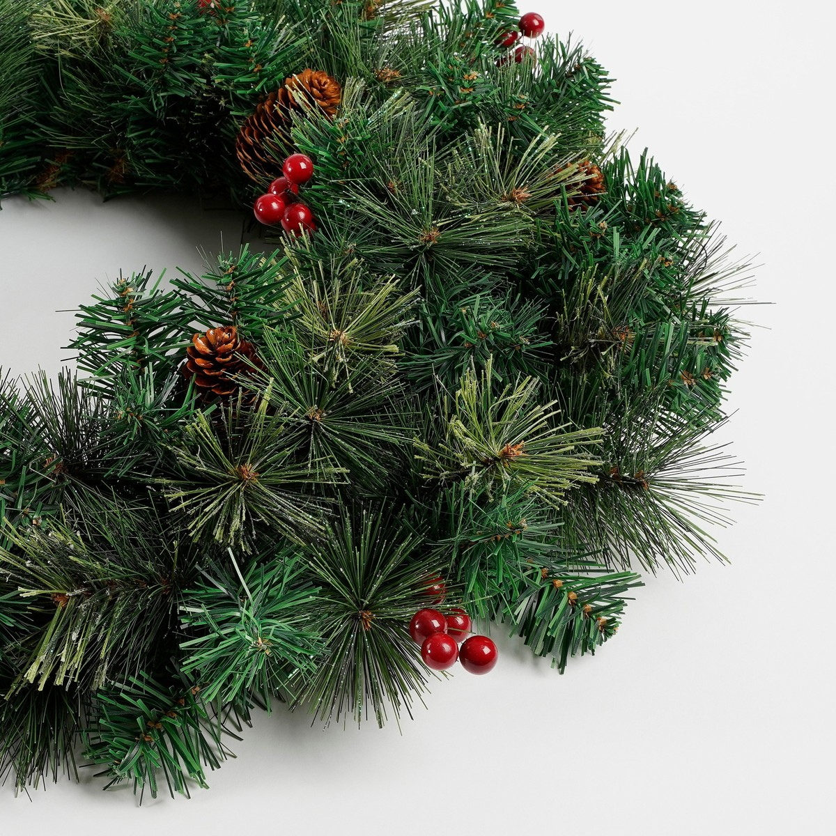 An artificial Christmas wreath of green pine branches decorated with pinecones and red berries on a white background.