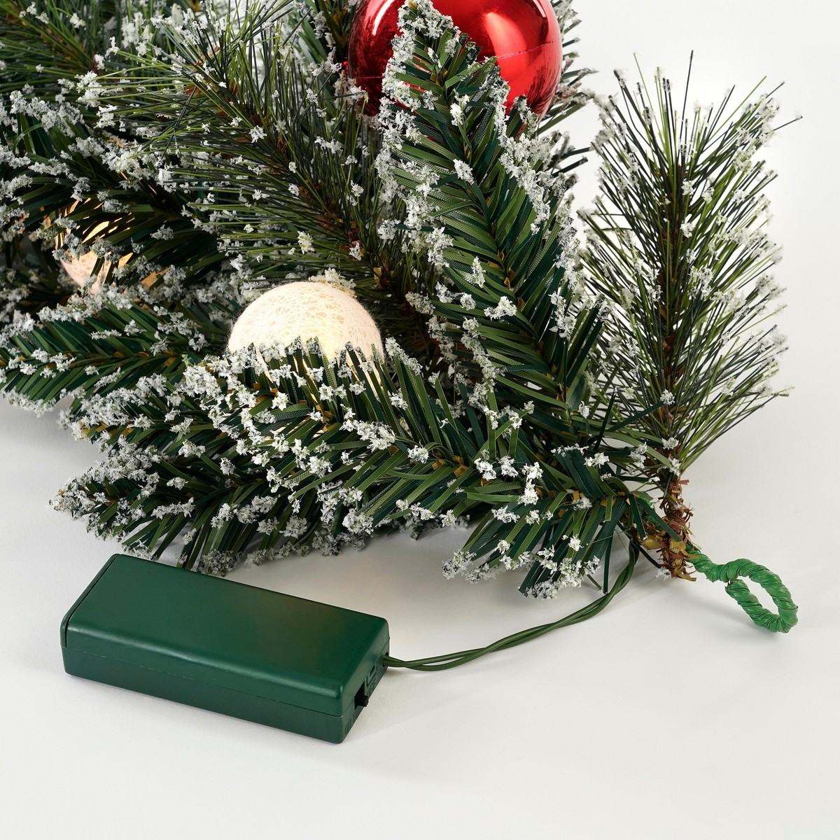 Verschneite künstliche Tannengirlande mit roter Christbaumkugel und Lichterkette an einer grünen Batteriebox.