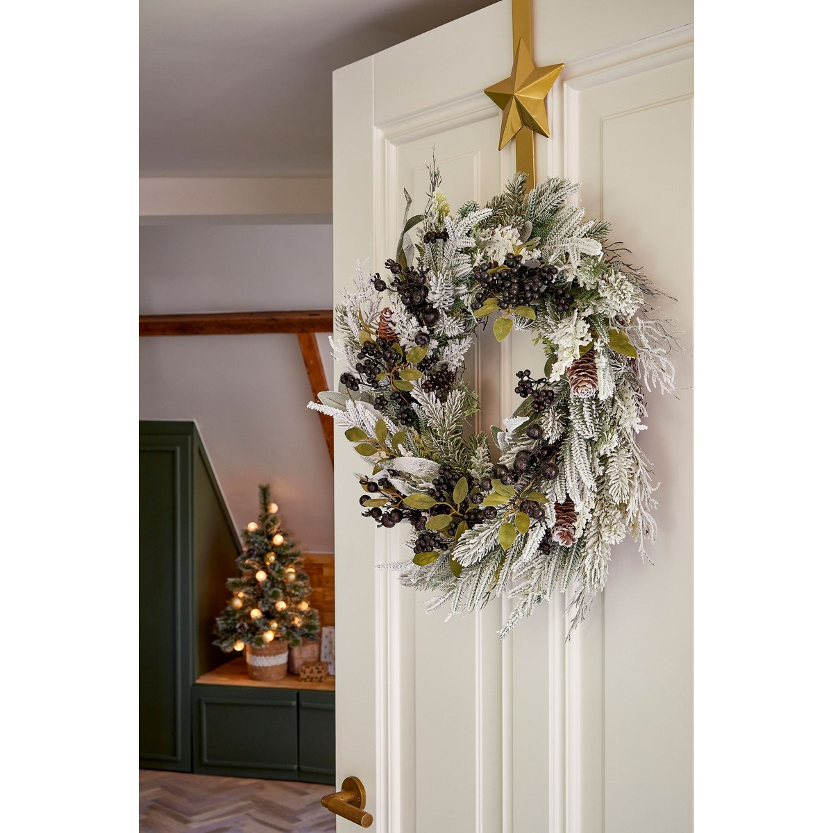 A snowy white Christmas wreath hangs on a white door beneath a golden star, with a small lit tree in the background.