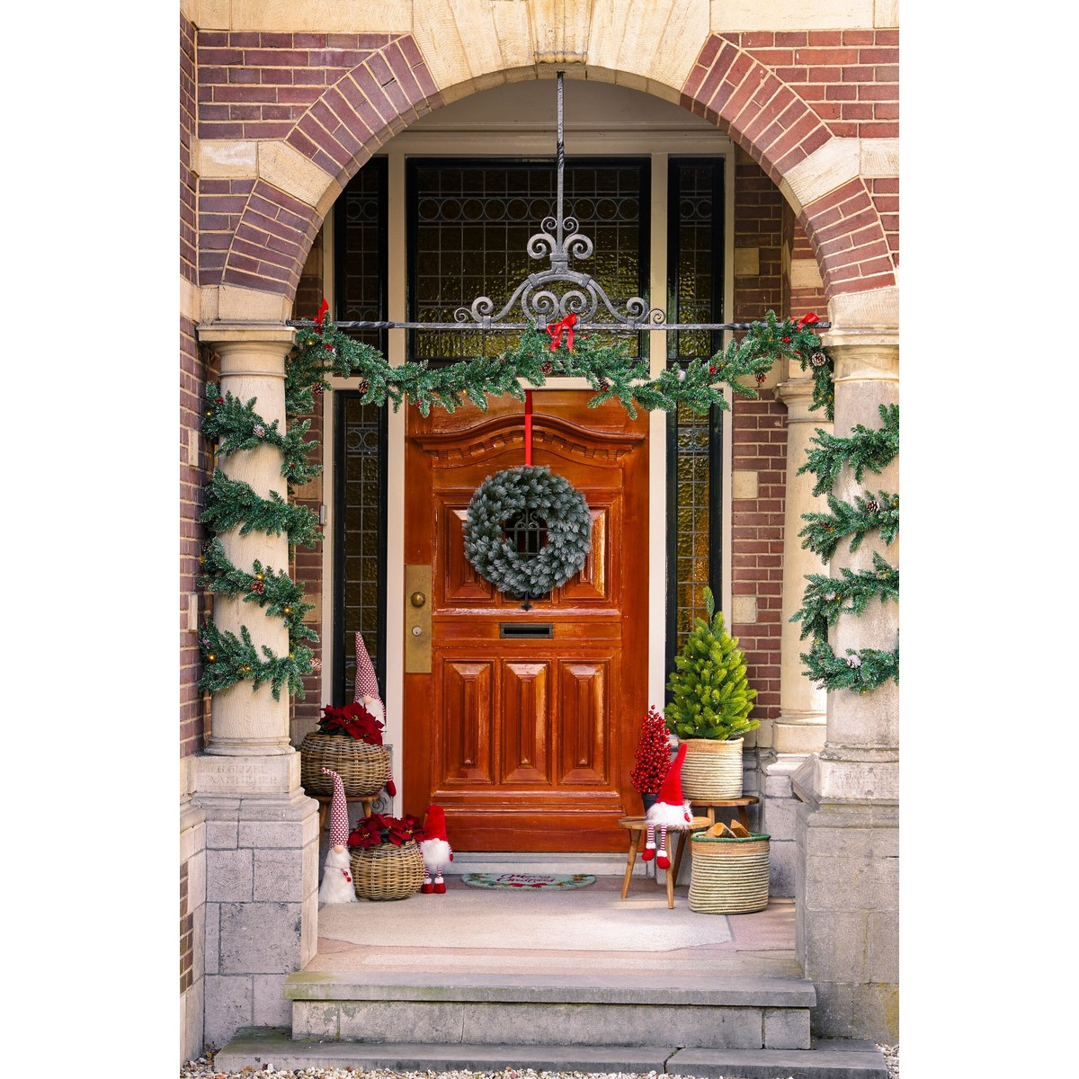 A wooden front door decorated for Christmas with a wreath, garlands, gnomes, and small trees in baskets under an arch.