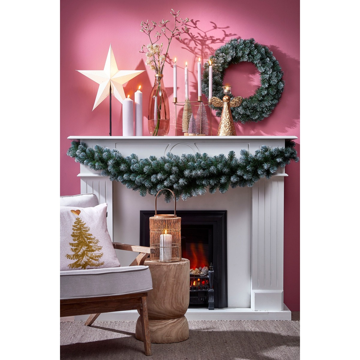 A white fireplace decorated with green garlands, candles, a glowing star, and a wreath against a pink wall.
