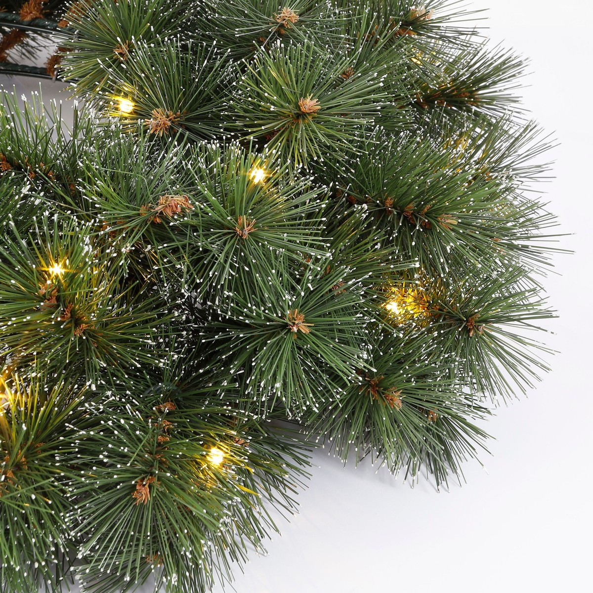 Close-up of a snowy green pine wreath decorated with warm glowing lights on a white background.