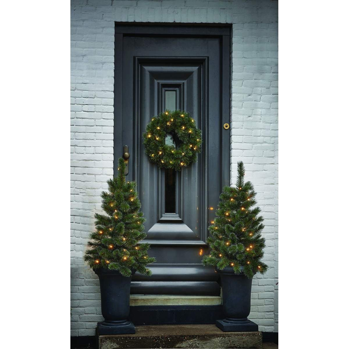 A black front door with a Christmas wreath and two lit potted pine trees in front.